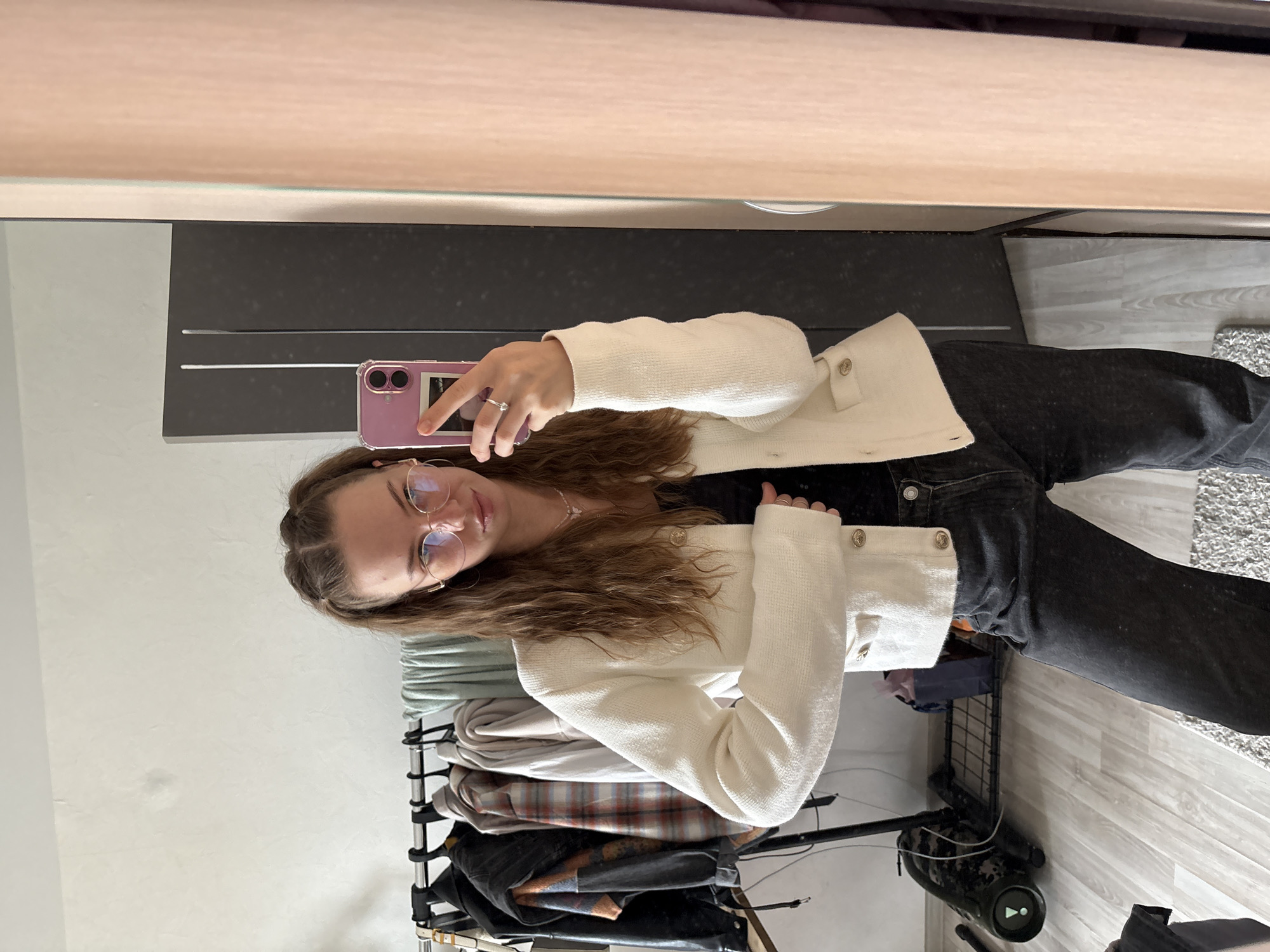 A young woman is taking a selfie in a bedroom. She is wearing a white tweed jacket, a black top, and dark wash jeans. She has long, wavy brown hair and is wearing round glasses. Her left hand is holding a pink phone, and her right hand is resting on her hip. In the background, there is a clothing rack with several items of clothing, a large black speaker, and a closet with wooden doors. The room has light-colored walls and light wood flooring, with a grey rug partially visible. The scene is indoors, likely in a residential setting, and there are no indicators of it being in the city of Perm, Russia.
