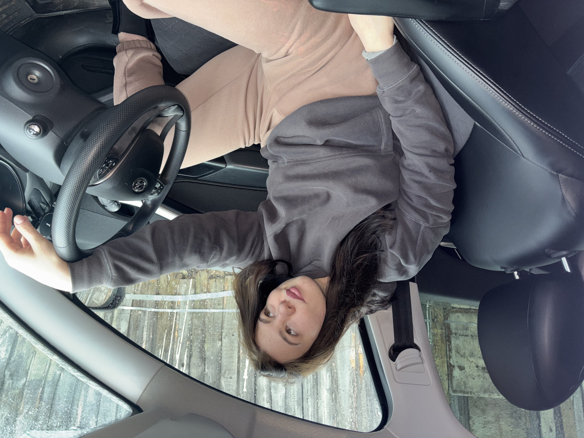 A woman is seated in the driver's seat of a car, facing the camera. She is wearing a grey long-sleeved hoodie and light brown sweatpants. The interior of the car is visible, including the steering wheel, dashboard, and passenger seat. Through the windshield, a view of what appears to be a rain-soaked window or fence is visible. The perspective of the image is inverted. The city and location are not discernible.