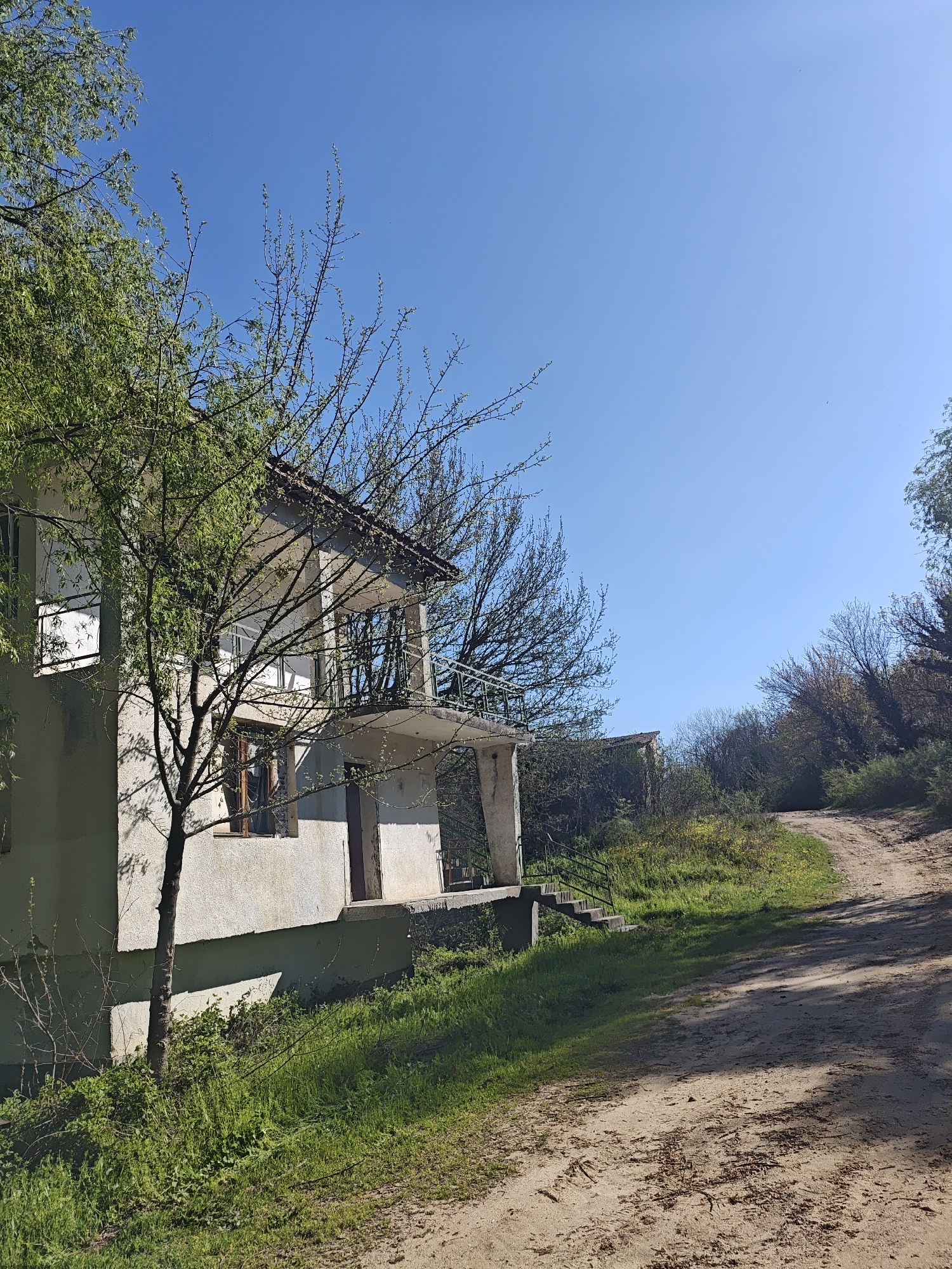 The image shows a two-story concrete house with a balcony. A tree is growing beside the house. The location appears to be rural or a very outer suburb, with a dirt road running alongside the house. The sky is clear and blue. There is another building further down the road. The image does not contain enough information to identify the city and state.