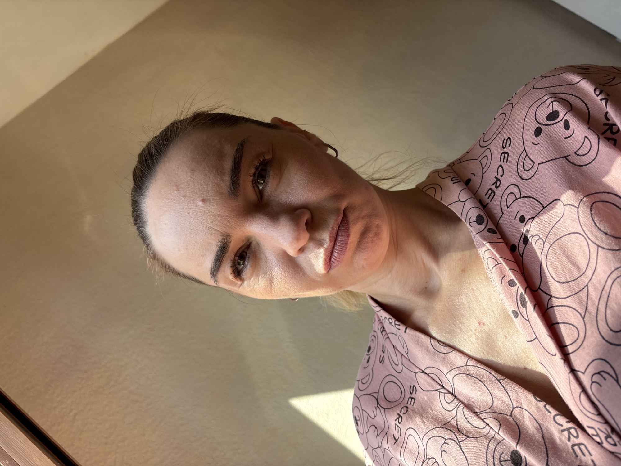 The media file is a close-up, portrait-style photograph of a woman. She is angled away from the camera, with her head tilted. She has dark eyebrows and appears to be wearing makeup, including lipstick. Her hair is pulled back. She is wearing a pink garment adorned with a repeating pattern of teddy bears and the word "SECRET" in black lettering. The lighting suggests it is daytime, with a soft light casting shadows on the wall behind her. The overall mood is neutral, with a hint of introspection. There is no discernible activity or event taking place beyond a posed photograph. The setting is indoors, with a plain, textured wall as the backdrop.