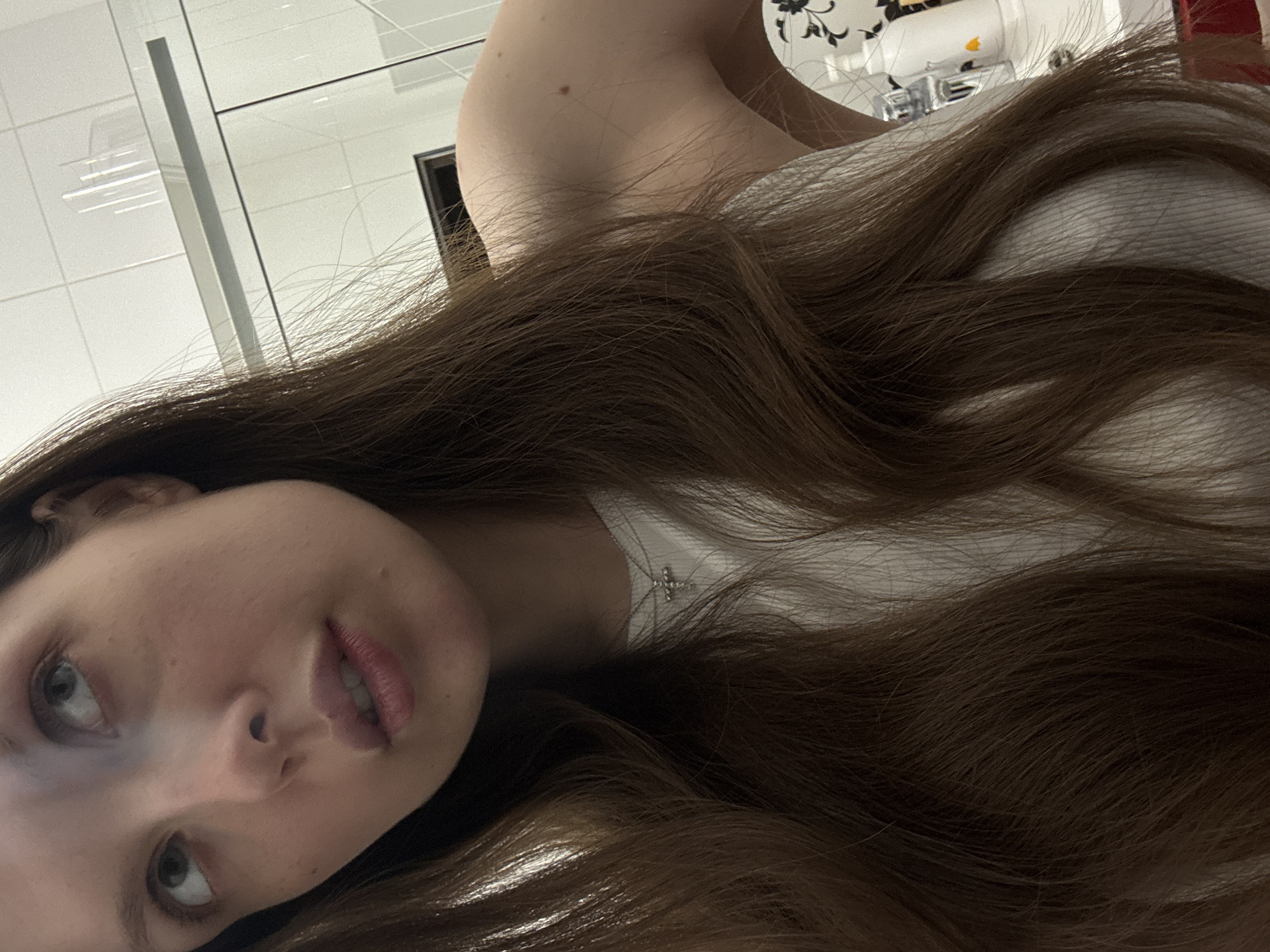 The image is an angled, close-up portrait of a young woman, likely a selfie, focusing on her head and upper torso. She has long, wavy, brown hair that dominates much of the frame, flowing towards the bottom and right. Her face, positioned in the lower left quadrant, is tilted, and she looks upwards and slightly to the left with light-colored eyes, which appear greenish-grey. Her lips are slightly parted. She is wearing a white top, and a delicate silver chain with a small cross pendant is visible against her skin.
The background indicates an indoor setting, possibly an office or commercial building, featuring white tiled walls and glass partitions. Reflections of overhead lights can be seen in the glass. In the upper right, there is a hint of a white object with black floral or leaf patterns, alongside what resembles a part of a sewing machine or similar mechanical apparatus. The lighting is artificial and bright. The woman's expression is neutral to slightly contemplative. The image was taken in Moscow, Russia. No visible text is present.