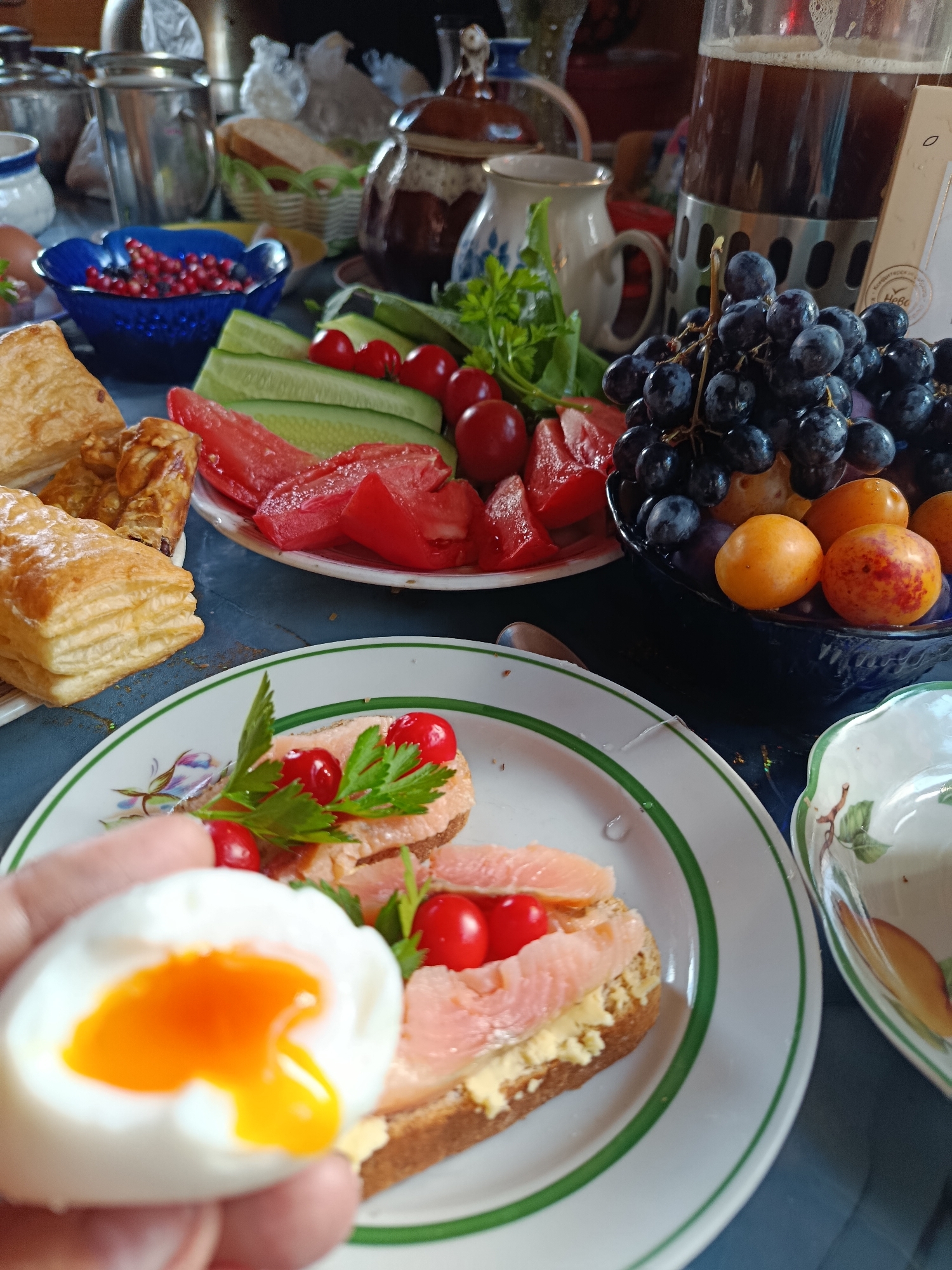 A table is laden with a breakfast spread, which includes a half-peeled soft-boiled egg held in a person's hand, revealing a runny, bright orange yolk. The egg is positioned in the foreground, slightly out of focus, with a plate of open-faced sandwiches topped with smoked salmon, small red berries, and a sprig of parsley in the middle ground. The sandwiches sit on a blue-patterned plate with a green rim. Behind the sandwiches, a platter holds sliced tomatoes and cucumbers. To the right, a dark blue bowl overflows with dark grapes and smaller orange fruits, likely plums or apricots. In the background, a large glass jug filled with a dark beverage, possibly beer or kvass, stands next to a rustic-looking ceramic teapot and other tableware. The setting appears to be indoors, on a table with a blue tablecloth, possibly in a home in Verea, Russia, given the overall rustic and traditional presentation of the food and tableware.
