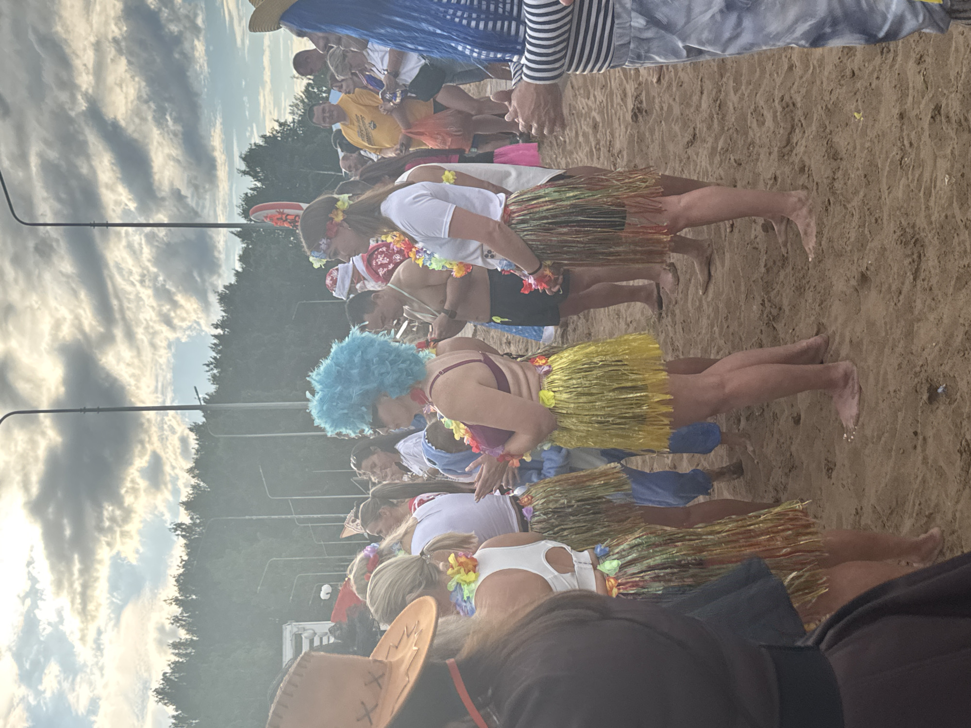 The image depicts a crowd of people on a sandy beach, likely in Chaikovsky, Russia, as indicated by the trees and streetlights common in the area. Many individuals are dressed in festive attire, including Hawaiian-themed outfits such as grass skirts and leis. Some are wearing brightly colored wigs. The sky is overcast with dramatic clouds. In the background, there are trees and several streetlights. The focus is on the group of people in the foreground, some of whom appear to be interacting or posing for photos. The overall atmosphere suggests a casual outdoor gathering or event.