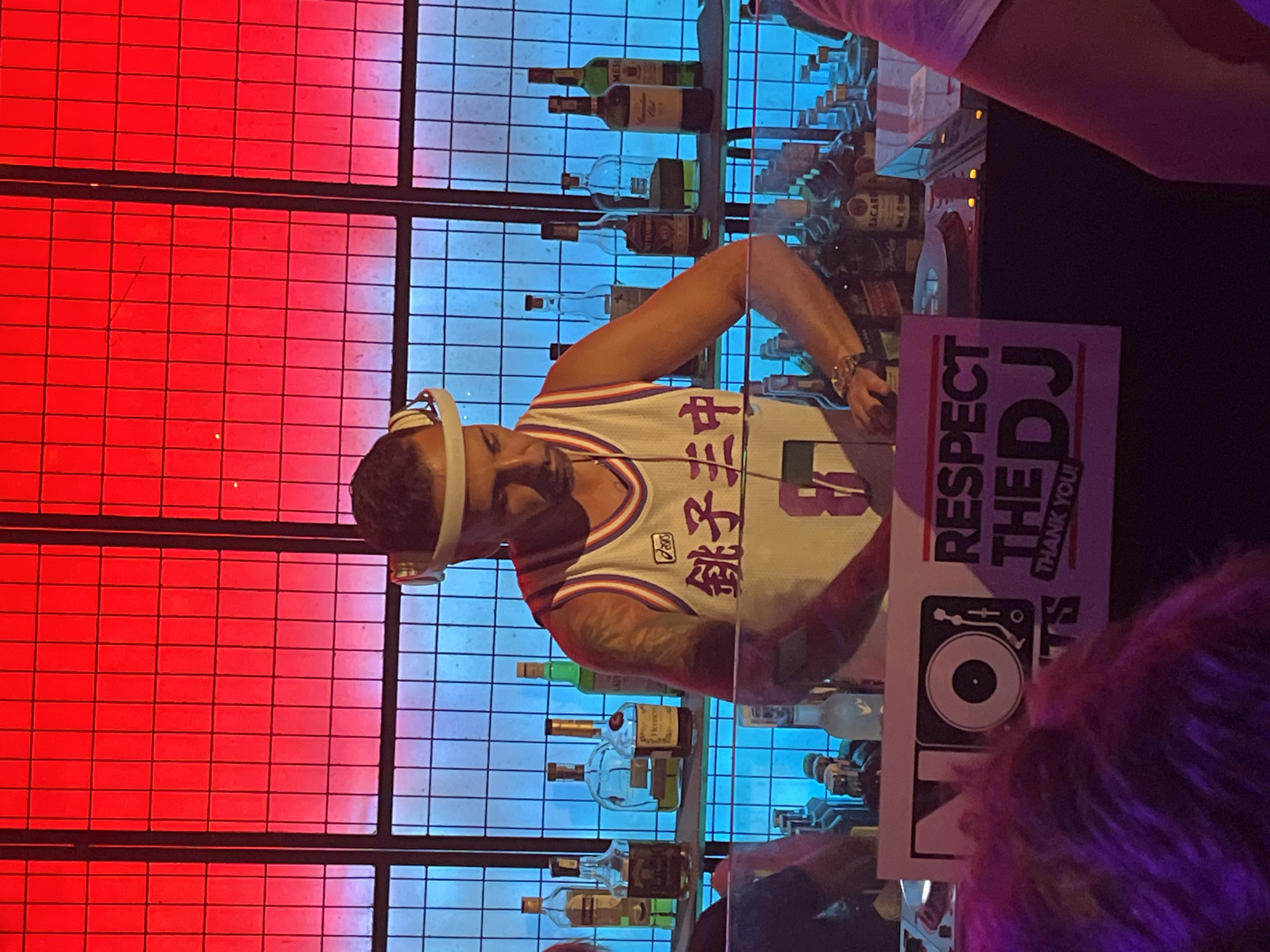 This full-shot image captures a male DJ actively performing in a dimly lit indoor venue, illuminated by vibrant red and blue neon lighting. The location in the city of Đà Nẵng, Vietnam, cannot be independently verified from the visual information presented.
The central figure is a man, appearing to be in his 30s or 40s, with short dark hair. He wears a white basketball-style sleeveless jersey with red and blue trim, featuring the "asics" logo and prominent purple/pink Japanese characters, "銚子王," above a number "8." White over-ear headphones are positioned around his head. A dark tattoo is visible on his right arm (viewer's left), and a watch is on his left wrist (viewer's right). His hands are engaged with the mixing equipment on a DJ booth in front of him.
Behind the DJ, a grid-like metal structure separates him from a bar setup. The upper portion of the background is bathed in a strong red light, gradually transitioning to a deep blue light in the lower section. Various liquor bottles, including Hennessy, Bacardi, Canadian Club, and Jameson, are arranged on shelves behind the DJ, illuminated by the blue light.
In the immediate foreground, a white sign is positioned on the DJ booth. It features a black square with a white vinyl record and turntable icon next to the word "NO" on the left, and the phrase "RESPECT THE DJ THANK YOU!" on the right. Blurred purple/magenta hair from an audience member is visible in the lower left corner, and a partial view of another person's arm in light-colored clothing is seen on the far right.
