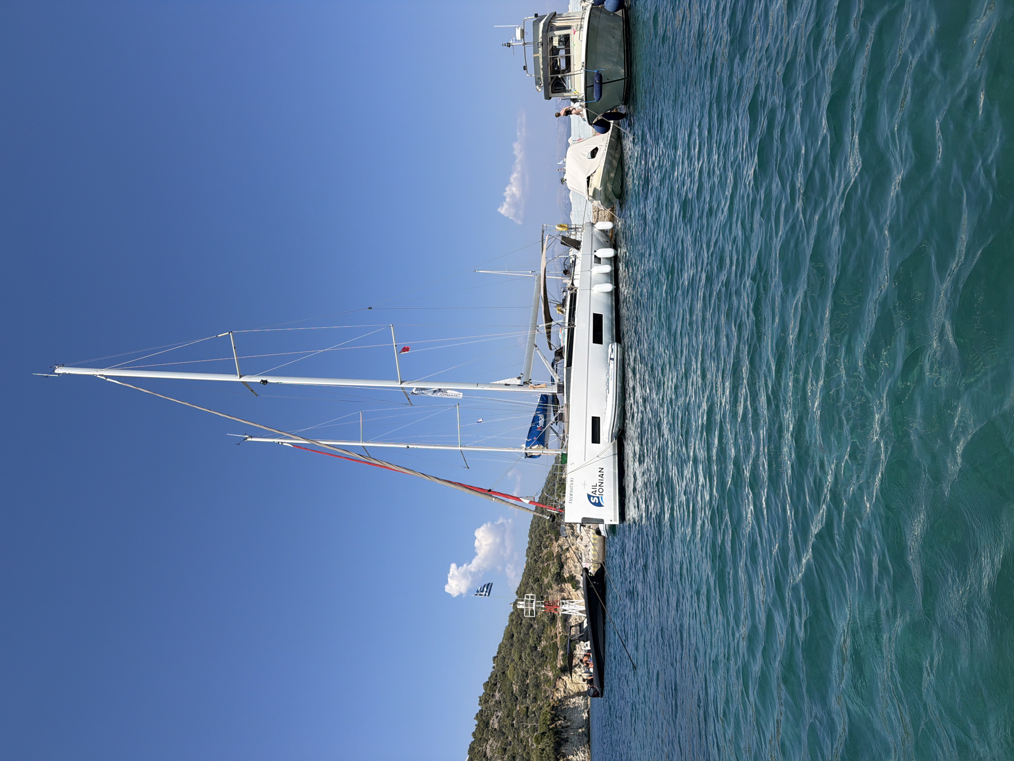 In the Municipal Unit of Kalamos, Greece, on a clear, sunny day with a bright blue sky, several boats are docked in a tranquil, turquoise body of water. A large white sailboat with a prominent mast dominates the center of the image, bearing the logo "SAIL IONIAN." To its left, a smaller dark-colored boat with several people aboard is tied to a buoy near a lighthouse. To the right of the sailboat, two smaller vessels are moored, one covered with a white tarp. Further to the right, a grey motorboat with a cabin is docked. The background features a lush, green, hilly coastline, suggesting a serene, coastal environment.