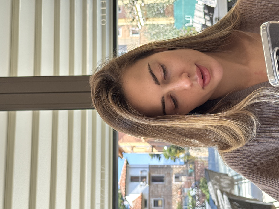 A female subject, with long light brown hair and a brown knitted top, is seated indoors, looking downwards at a smartphone held in her right hand, partially visible at the bottom right. Her eyes are directed towards the phone, and her lips are slightly parted. Behind her, a large window reveals an outdoor scene in Albisano, Italy. Outside, blurred outdoor seating, including chairs and tables, is visible. Beyond this, traditional two-story buildings with light-colored facades and orange-tiled roofs are present, one featuring a balcony. Green foliage is also visible. The sky is clear, suggesting daytime. Horizontal blinds are visible in the upper portion of the window, and the reversed text "OTTELLO" is discernible on the glass.