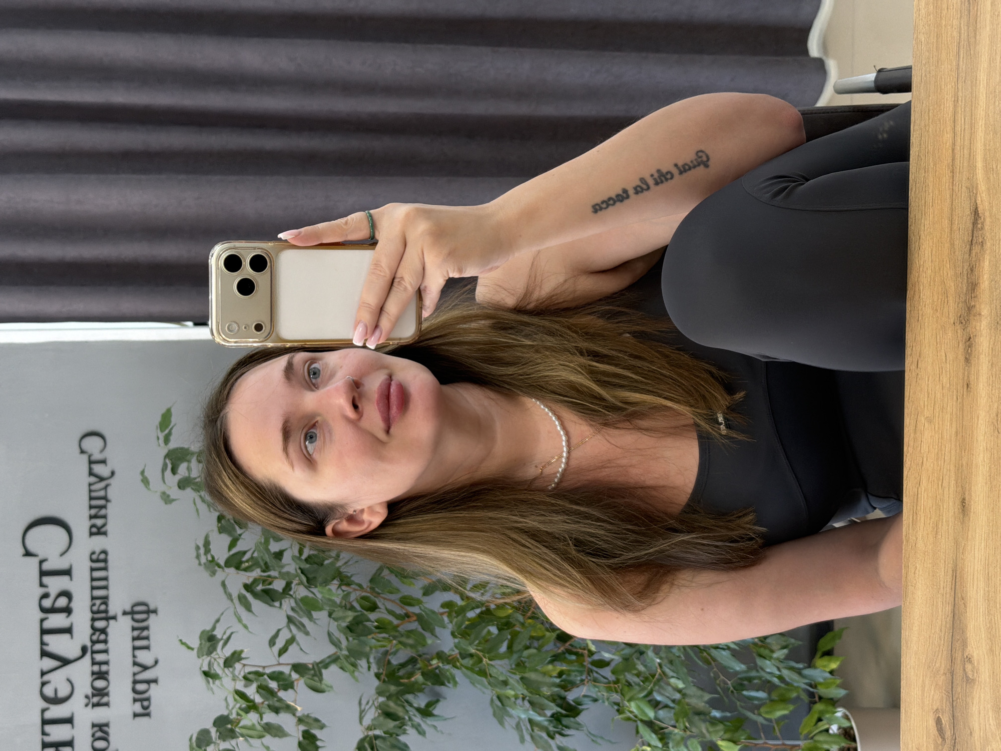 A young adult female, with long light-brown hair and blue eyes, is seated indoors in what appears to be a permanent makeup studio in Chaikovsky, Russia. She is taking a selfie with a gold-framed smartphone, held in her right hand, while looking slightly to her right with a subtle smile. She wears a black athletic top and dark grey leggings. A string of pearls and a thin gold-colored chain are visible around her neck. A thin dark-green ring is on her right ring finger. Her inner left forearm displays a vertical black text tattoo, which is partially obscured and mirrored due to the selfie angle. Her right ear has a small silver stud in the helix, and a silver stud is present on her left nostril.
The background features a light grey wall with mirrored, backward black Russian text that, when correctly oriented, reads "СТУДИЯ ПЕРМАНЕНТНОГО МАКИЯЖА" (PERMANENT MAKEUP STUDIO). A tall, leafy green potted plant stands to her left. To her right, a dark grey, pleated curtain is visible. A wooden-textured surface, likely a desk or table, spans the bottom of the foreground.