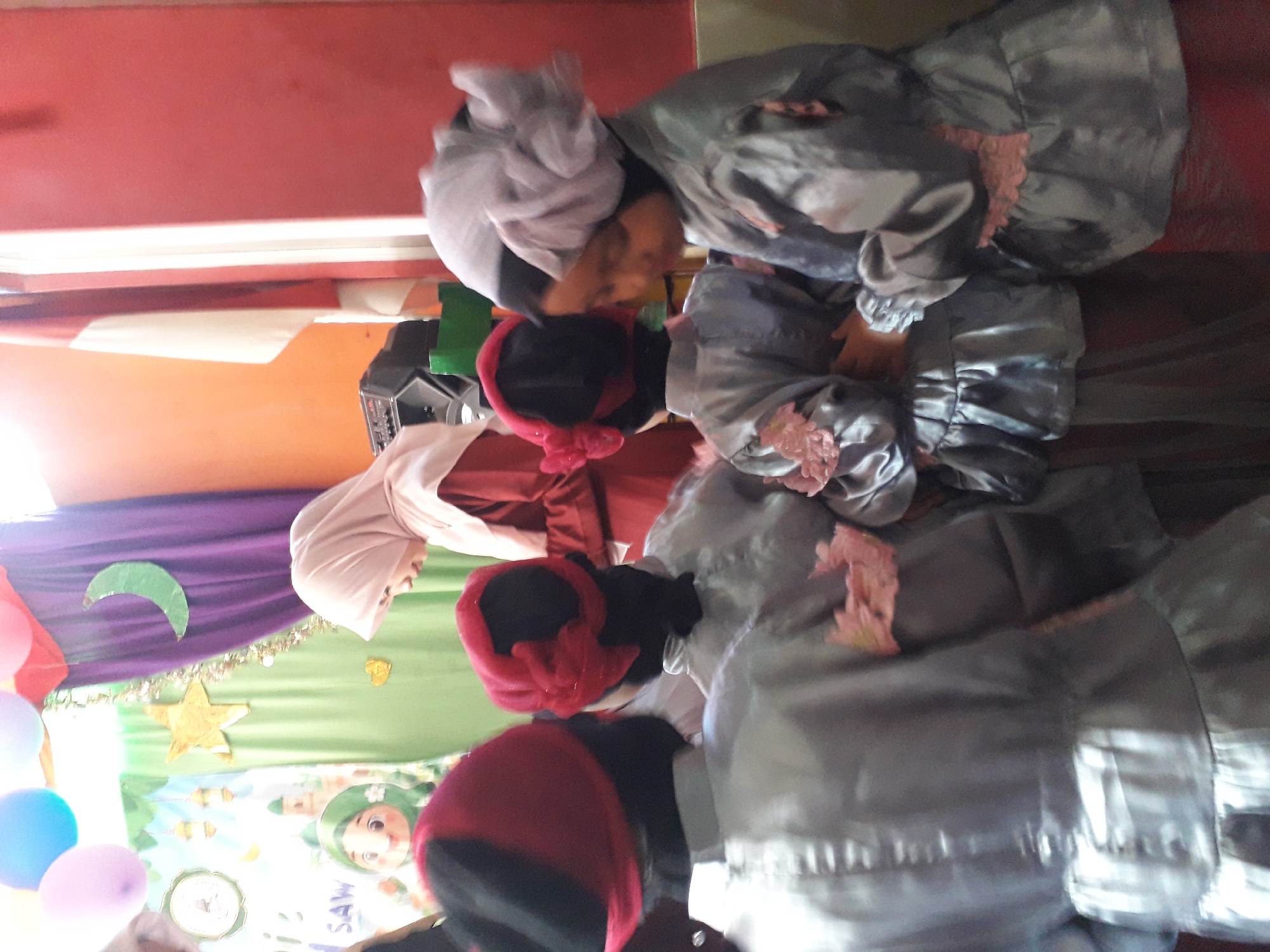 The image depicts an indoor scene, possibly a classroom or event space, with a colorful backdrop of green and purple curtains, balloons, a crescent moon cutout, and a gold star. A woman wearing a light pink hijab and a red dress stands in the background, facing downwards, possibly addressing a group. In the foreground, four children, dressed in matching silver outfits with red head coverings adorned with ribbons or bows, are partially visible from behind. Their attire suggests they may be participating in a cultural or religious event. The children's faces are not clearly visible. To the right of the woman in the background, a flag with red and white stripes is hung vertically. The overall atmosphere appears festive and celebratory, possibly related to an Islamic holiday given the crescent moon and children's attire. The exact location within Cankuang, Indonesia, cannot be determined from the image alone.