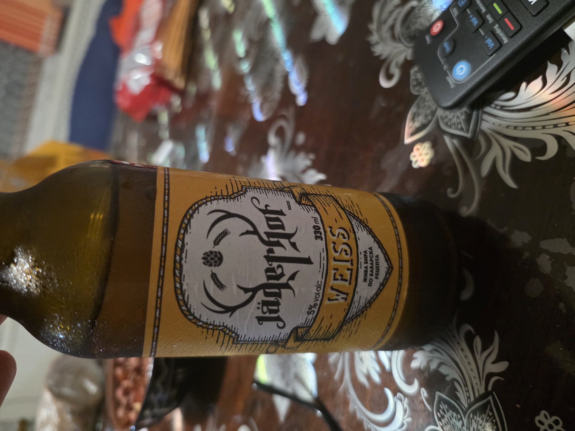 The image depicts a close-up view of a bottle of "Jägerhof" Weiss beer. The label is prominently displayed, featuring a stag's head with antlers, a hop cone, and text indicating "5% vol alc" and "330 ml". Below this, the word "WEISS" is in large letters, followed by Cyrillic text that reads "ЖИВА БИРА ПО БАВАРСКА РЕЦЕПТА" which translates to "Live beer according to Bavarian recipe." The bottle is frosted with condensation, suggesting it is chilled.
The bottle is placed on a dark, patterned surface that appears to be a table. In the background, slightly out of focus, are some items including a bowl of what looks like nuts or food, and some red and white packaged goods. To the right of the bottle, a black remote control is visible, with several buttons including power, volume controls, and recording functions. The overall lighting suggests an indoor setting, possibly a home or restaurant.
There is no information or visual evidence in the image that would allow for the identification of the specific location within the city of Targovishte, Bulgaria. The image solely focuses on the beer bottle and its immediate surroundings on a table.