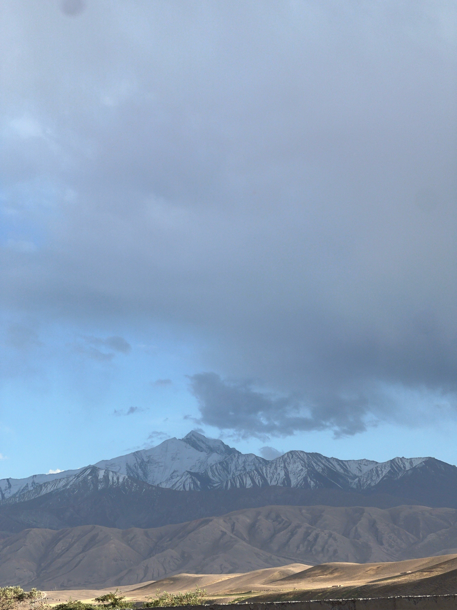 A wide, vertical shot captures a vast mountain range under a cloudy sky in Daroot Korgon, Kyrgyzstan. The sky is predominantly filled with gray and white clouds, with patches of blue sky visible towards the horizon. The top portion of the image is entirely dominated by these clouds, creating a diffused light. Below the clouds, a prominent snow-capped mountain range stretches across the frame. The peaks are jagged and rugged, with snow covering the upper portions. In front of the snow-capped mountains, a series of rolling, barren hills are visible. These hills have a dusty, earthy tone, with shadows and highlights indicating their undulating forms. The foreground shows a dry, scrubby landscape with a low wall or barrier in the very bottom. The overall scene conveys a sense of rugged, natural beauty in a remote, mountainous region.