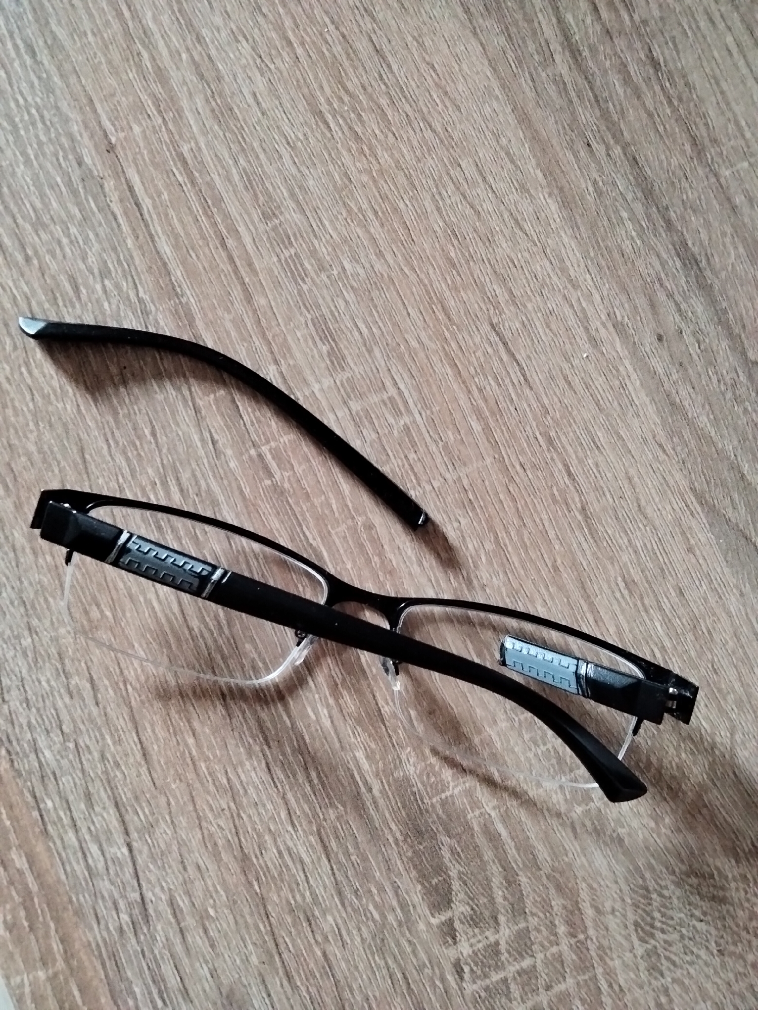 A pair of broken black eyeglasses lies on a light brown wooden surface. The eyeglasses are fragmented, with one arm detached from the main frame. The frame is thin and appears to be made of metal, with a decorative element on the visible temple. The wooden surface has a visible grain pattern. The image is taken from a top-down perspective. There is no indication of the location being Kyiv, Ukraine.