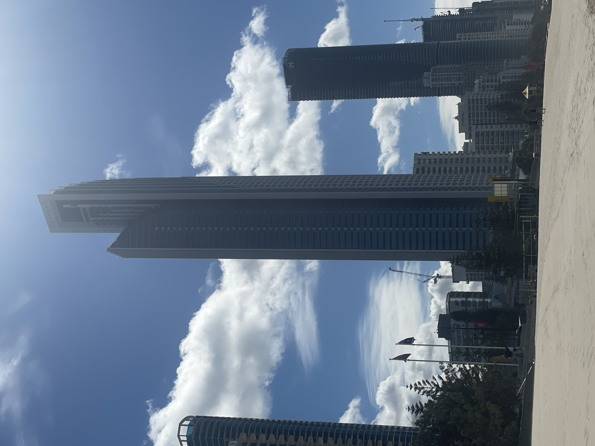 The image captures a view of the Surfers Paradise skyline in Australia, facing inland from a sandy beach under a partly cloudy sky. Dominating the center is the tall, slender skyscraper "Peppers" with its name visible at the top. To its left, another skyscraper with a rounded facade and a balcony structure is partially visible. To the right of Peppers, a dark, rectangular skyscraper stands, and further to the right, a series of other high-rise buildings form a dense urban backdrop. Several flags are planted on poles on the left side of the image, and a construction crane is visible among the buildings, suggesting ongoing development. In the foreground, the sandy beach is empty except for a few scattered individuals and beach infrastructure like a volleyball net. The scene depicts a bright, sunny day with scattered white clouds against a blue sky.