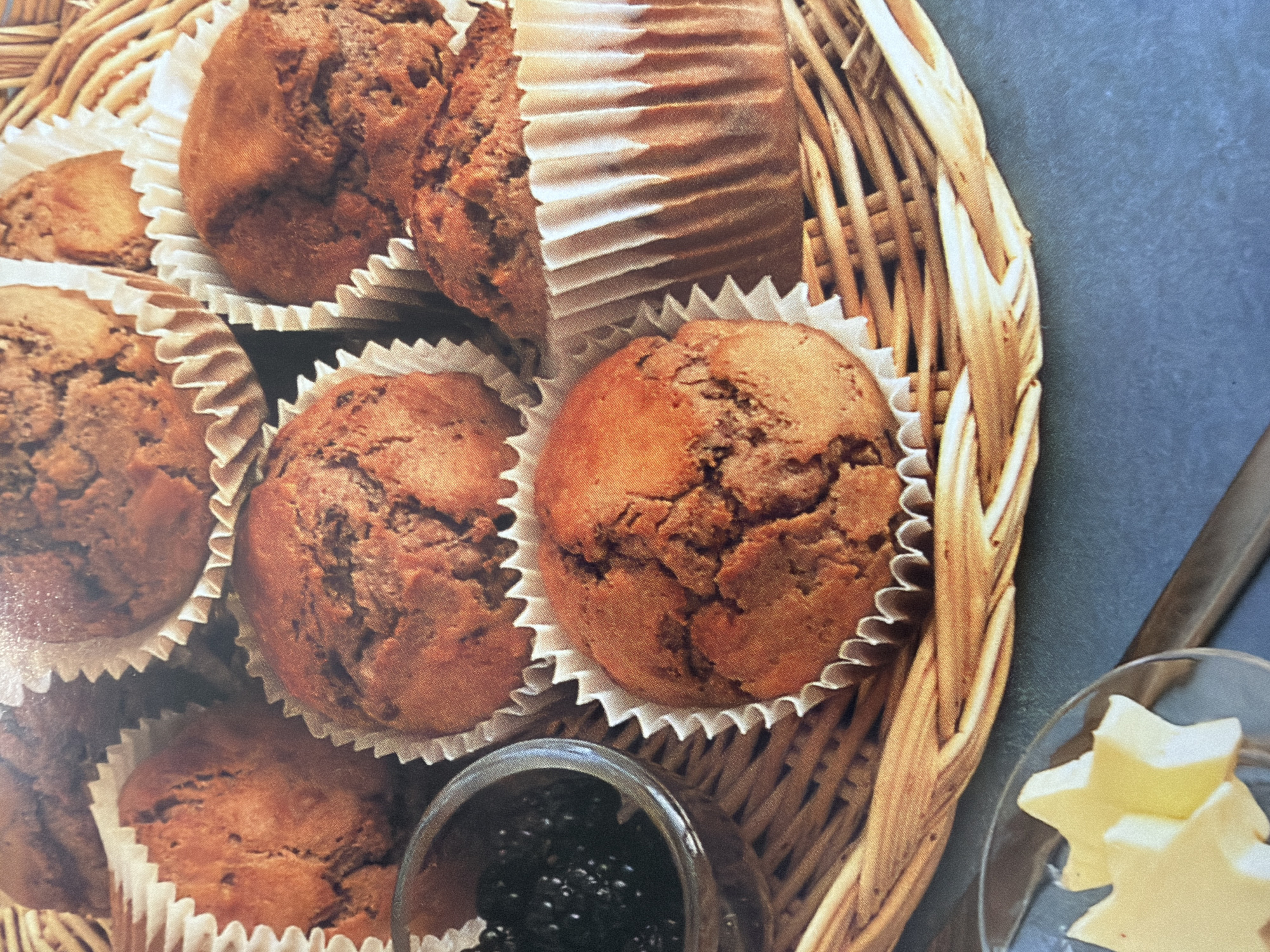A close-up, overhead shot shows a woven basket filled with chocolate muffins. The muffins are in paper liners and some are stacked. To the left of the basket, a small glass bowl contains dark jam. Below the basket, a small glass dish holds two star-shaped pieces of butter. The background is a dark, textured surface. The scene is set in an unknown location.