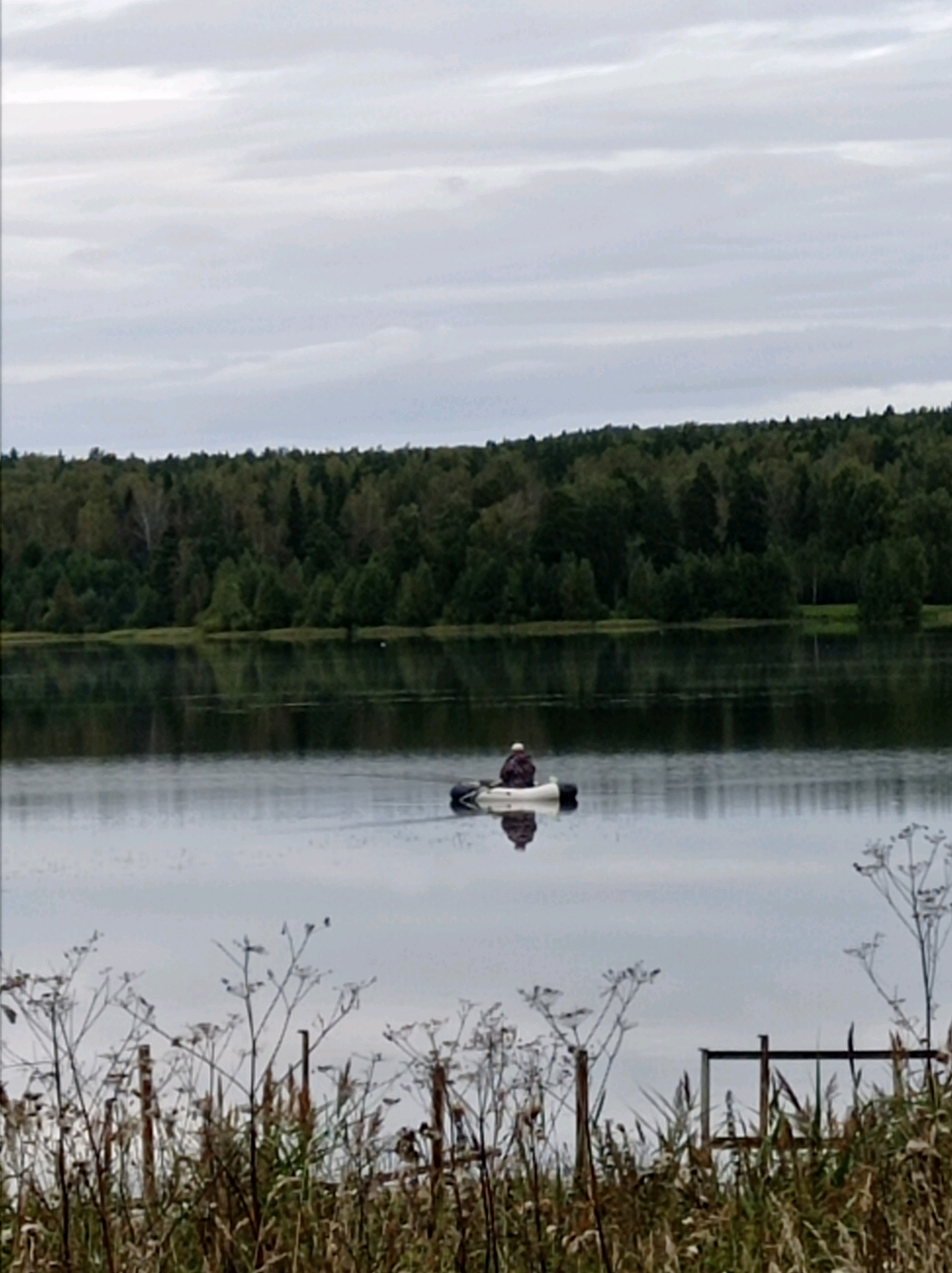 A person in a small inflatable boat is on a calm lake. The person is wearing a dark, patterned jacket and a cap. The boat is white with black tubes. The lake is surrounded by a dense forest of green trees. The sky is overcast with gray clouds. In the foreground, dry plants and reeds are visible, and a wooden fence can be seen in the lower right corner. This scene is likely located in or near the city of Degtyarsk, Russia, given the context of the user's request.