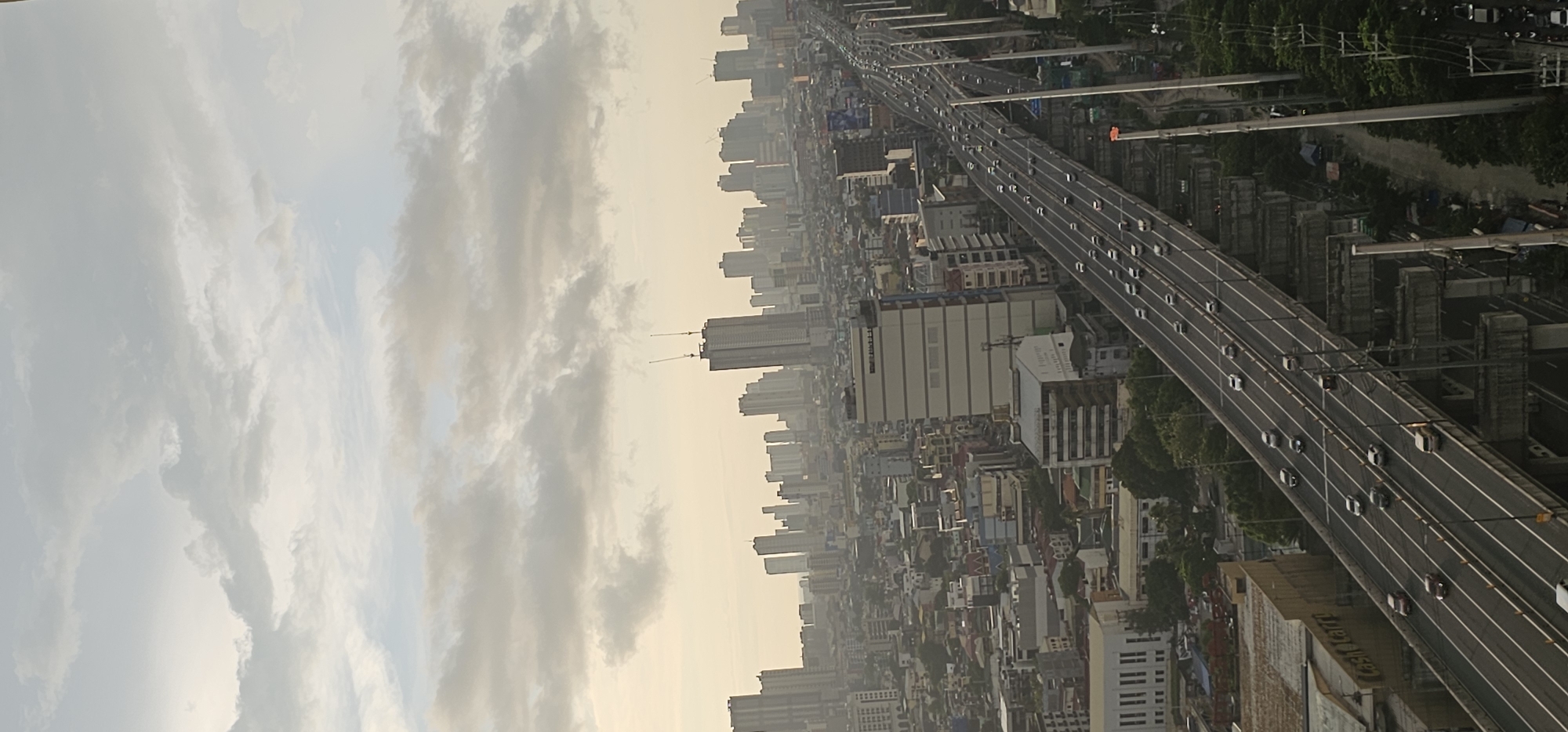 The image captures a panoramic view of the city of Makati, Philippines, under a cloudy sky. In the foreground, a multi-level elevated highway is visible, with numerous cars traveling in both directions. The highway is a dominant feature, constructed with concrete pillars and a paved road surface. Below the highway, the urban landscape unfolds, characterized by a dense collection of buildings of varying heights and architectural styles. Commercial buildings, residential blocks, and what appear to be office towers make up the cityscape. Two tall buildings under construction, with cranes atop them, stand out in the mid-ground, indicating ongoing development. The buildings are mostly concrete and glass structures, reflecting the overcast sky. The overall impression is one of a bustling metropolitan area, with a vast network of roads and a high density of buildings. The light suggests it might be late afternoon or early morning, with the sun diffused by the clouds.