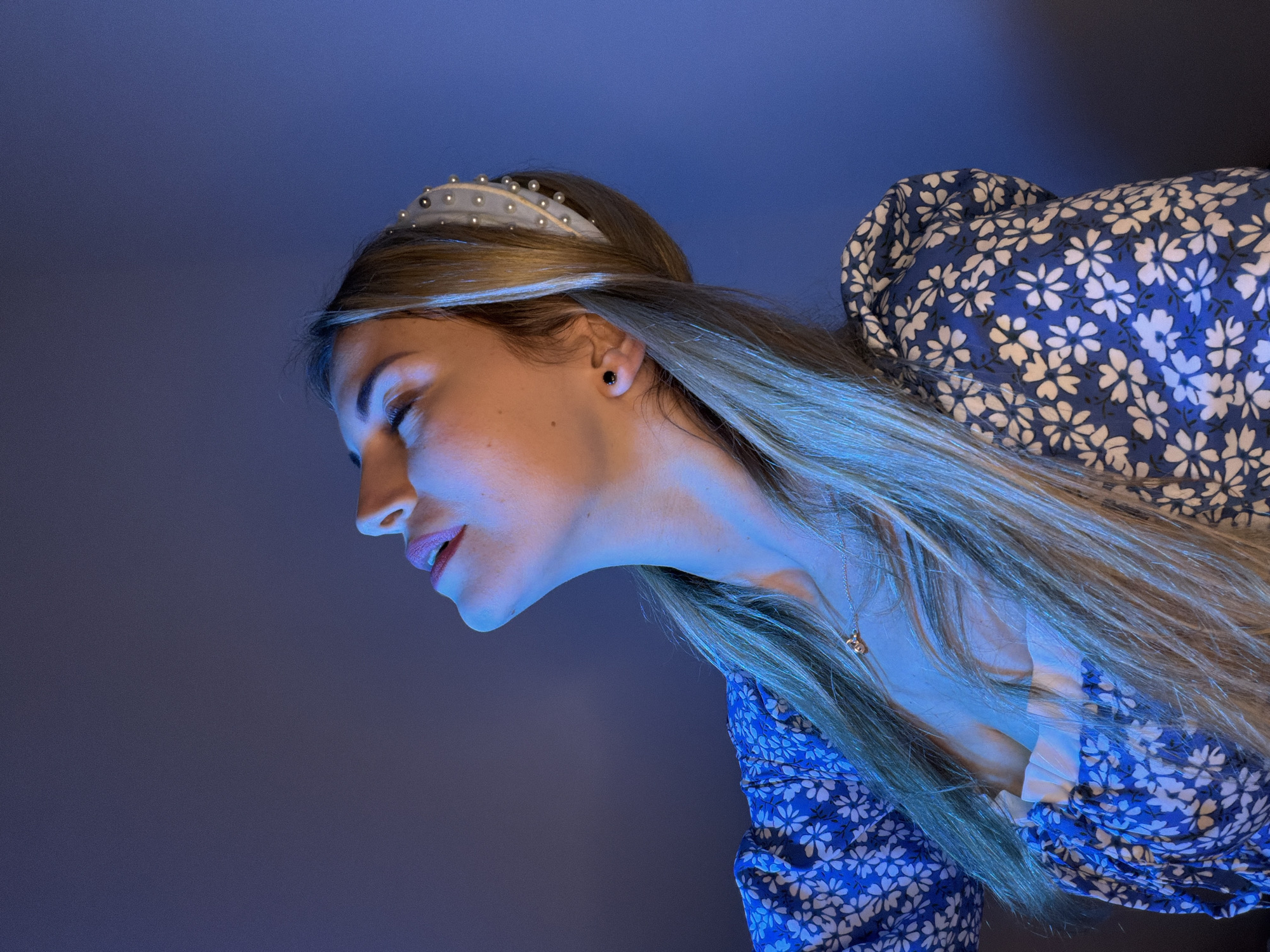 A young woman with blonde hair wearing a pearl-studded headband and a blue floral dress is angled towards the bottom left of the frame. Her eyes are closed, and she appears to be looking down. She is wearing a necklace with a small pendant. The background is a gradient of blue and gray. The location and the city of Budva, Montenegro, are not discernible from the image.
