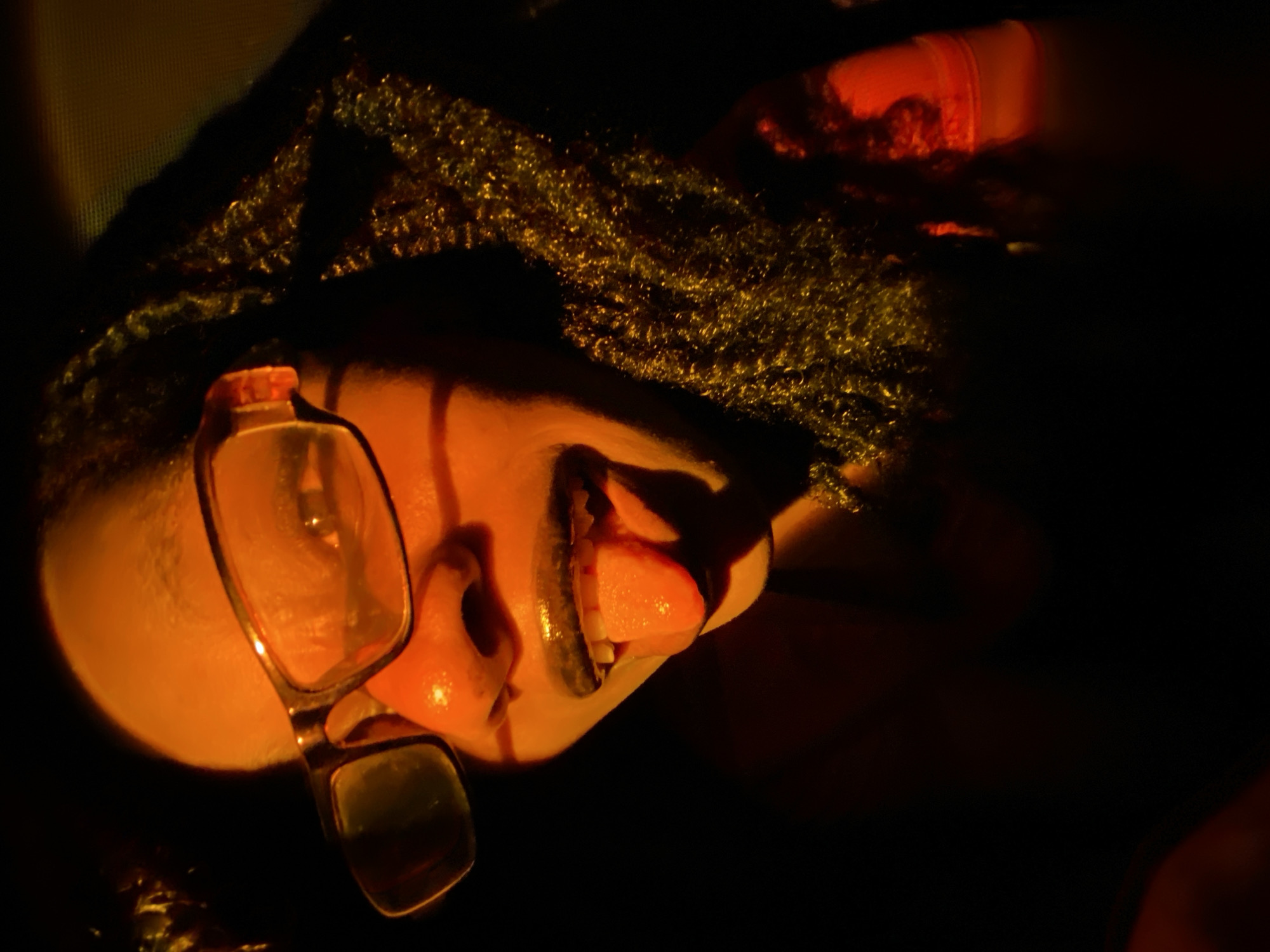 A close-up, low-angle shot shows a person's face tilted upwards, illuminated with warm, orange lighting. The person is wearing glasses with a clear frame, and their mouth is open, revealing their tongue. The top of their head is visible, adorned with what appears to be a textured head covering, possibly hair or a wig. The background is predominantly dark, creating a strong contrast with the illuminated face. A hint of orange clothing is visible on the right. The location is unknown.