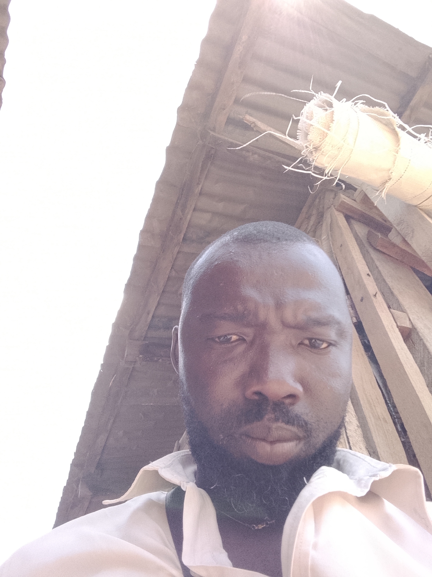A low-angle selfie shows a Black man with a shaved head and a dark beard, looking directly at the camera with a neutral expression. He wears a light-colored shirt and a dark strap is visible over his shoulder.
Behind him is a partially constructed structure, possibly a building under renovation or construction, evidenced by wooden beams and a corrugated metal roof. A cylindrical object wrapped in fibrous material is attached to the structure.
Given the high intensity of the light and the partially built structure, the location is likely outdoors in a developing urban or peri-urban environment. Without additional context, it is impossible to confirm the precise location in Jos, Nigeria.