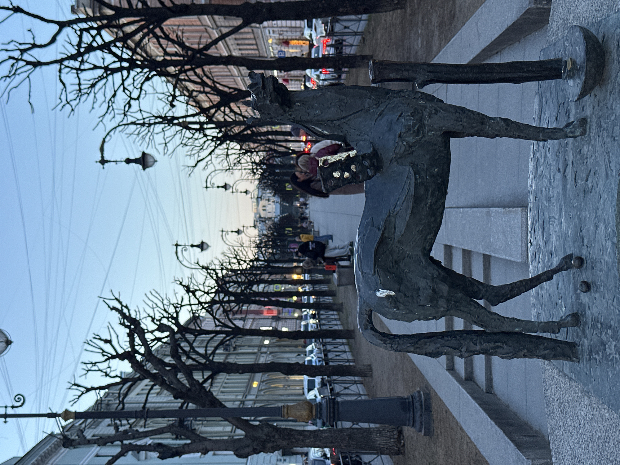 The image captures a bronze sculpture of a dog known as Gavryusha, located in Saint Petersburg, Russia. The sculpture is positioned on a low pedestal along a street with a row of bare trees and period-style street lamps. In the background, there is a pedestrian walkway with people, and further back, there are buildings and moving vehicles. A person is interacting with the sculpture, possibly touching or photographing it. The sky is a pale, muted blue.