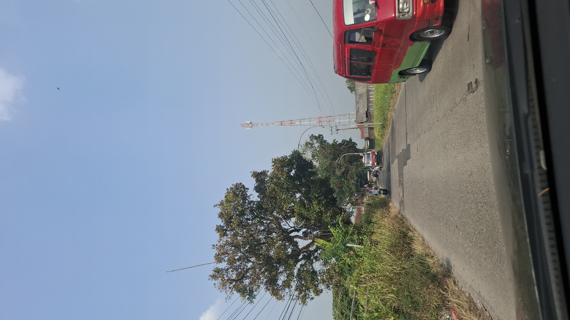 The image captures a view from a vehicle traveling on a paved road in Sukasari, Indonesia. The road is lined with dense vegetation on the left, including tall grasses and a large, leafy tree with some brown foliage. On the right side of the road, a red and green van is visible. Further down the road, a cell tower stands tall against a clear blue sky. Other vehicles, including a white truck and several motorcycles, are also present on the road, suggesting moderate traffic. The scene appears to be in a rural or semi-rural area, with a few buildings visible in the distance. The overall lighting indicates it is daytime, likely mid-morning or early afternoon, with sunlight casting shadows.