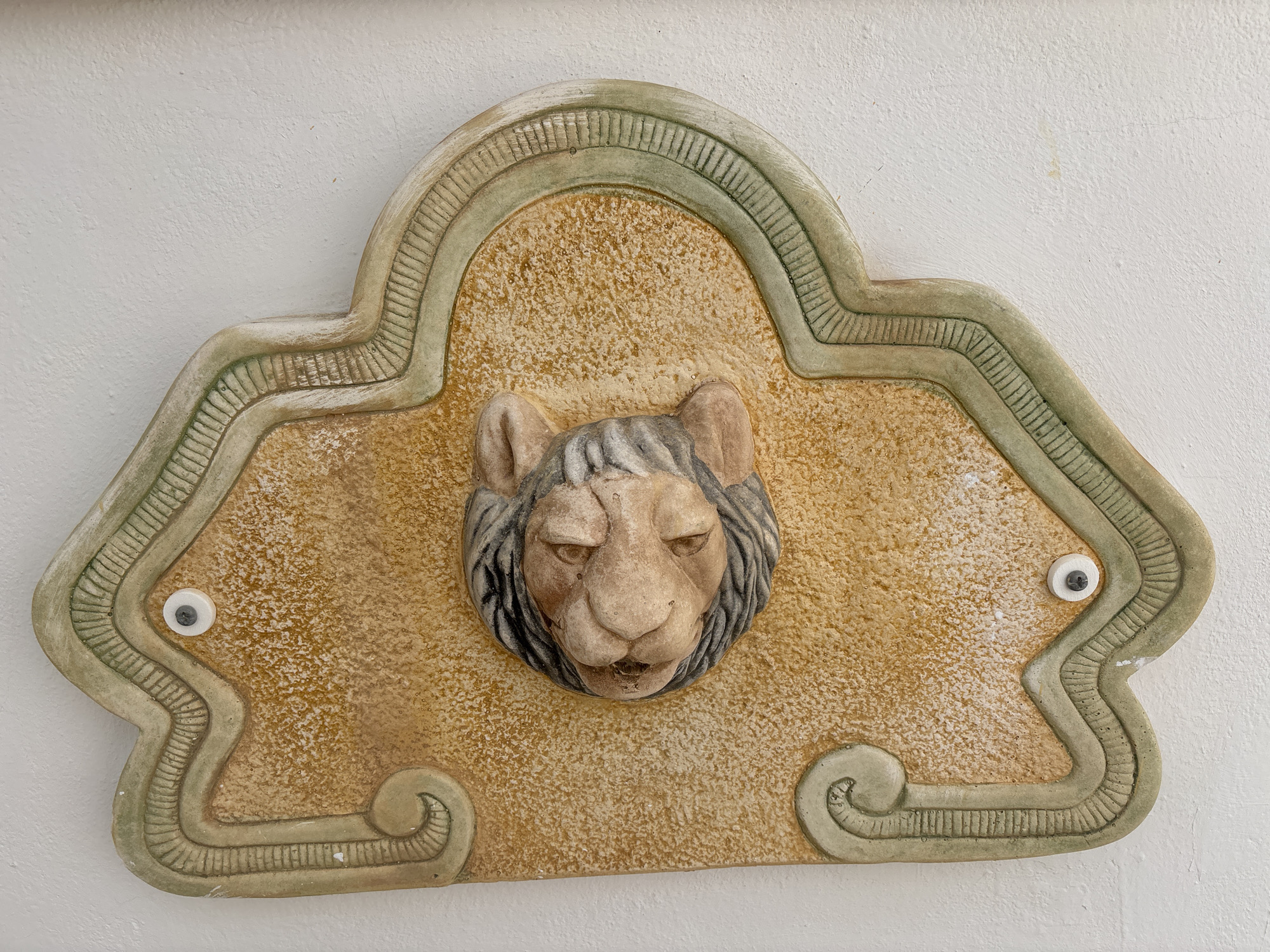 The image shows a close-up, eye-level shot of a decorative plaster relief mounted on a white textured wall. The relief is a stylized, somewhat weathered depiction of a lion's head with a mane, set against a textured ochre background. The lion's face is rendered with a scowling expression, with its mouth slightly open. The relief is framed by an ornate border in muted green and beige tones, featuring raised ribbed patterns and scrollwork. Two small, googly-eye-like ornaments, one on each side of the relief, are visible, adding a whimsical touch. This decorative element is likely part of the exterior facade of a building in the city of Orihuela, Spain, though its specific location within the city cannot be determined from the image alone.