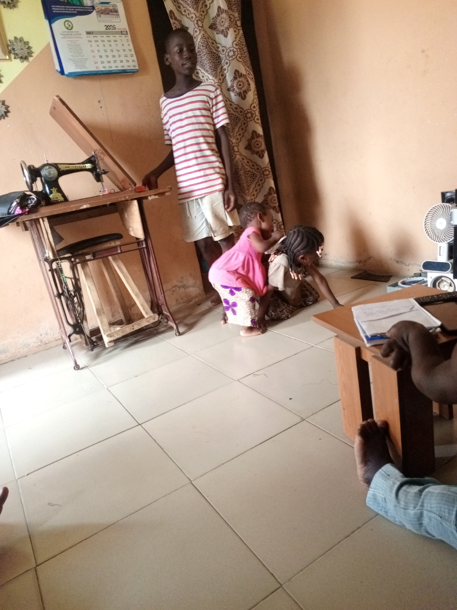Here's a detailed description of the image:
**Setting:**
The photo appears to be taken in an interior space, likely a room within a residence. The walls are painted in a light peach color. The floor is covered in light-colored square tiles. The light source seems to be a combination of natural and artificial light.
**People:**
* **Older Boy:** A young boy, wearing a white and red striped shirt and light-colored shorts, is standing near a sewing machine table.
* **Two Younger Children:** Two young girls are present. One is wearing a pink top and a patterned skirt and appears to be attempting to climb on the back of the other, who is crawling on the floor. The crawling child is wearing a light-colored top and dark skirt.
* **Person Seated:** Part of a person is visible in the lower right corner, showing a hand and a foot wearing a blue shirt.
**Objects:**
* **Sewing Machine:** A vintage sewing machine is positioned on an old-fashioned table.
* **Calendar:** There's a calendar hanging on the wall, with "2026" prominently displayed.
* **Curtain:** A patterned curtain is hanging near the wall.
* **Small Table:** There is a small brown table with papers and some electronic devices on it.
* **Fan:** A white electric fan is visible behind the table.
**Actions and Interactions:**
* The older boy is standing near the sewing machine.
* The two younger children are interacting playfully, with one attempting to climb on the back of the other.
**Environment:**
The room appears to be a living space, with the presence of domestic items. The tiled floor and the color of the walls indicate a relatively simple and modest setting.