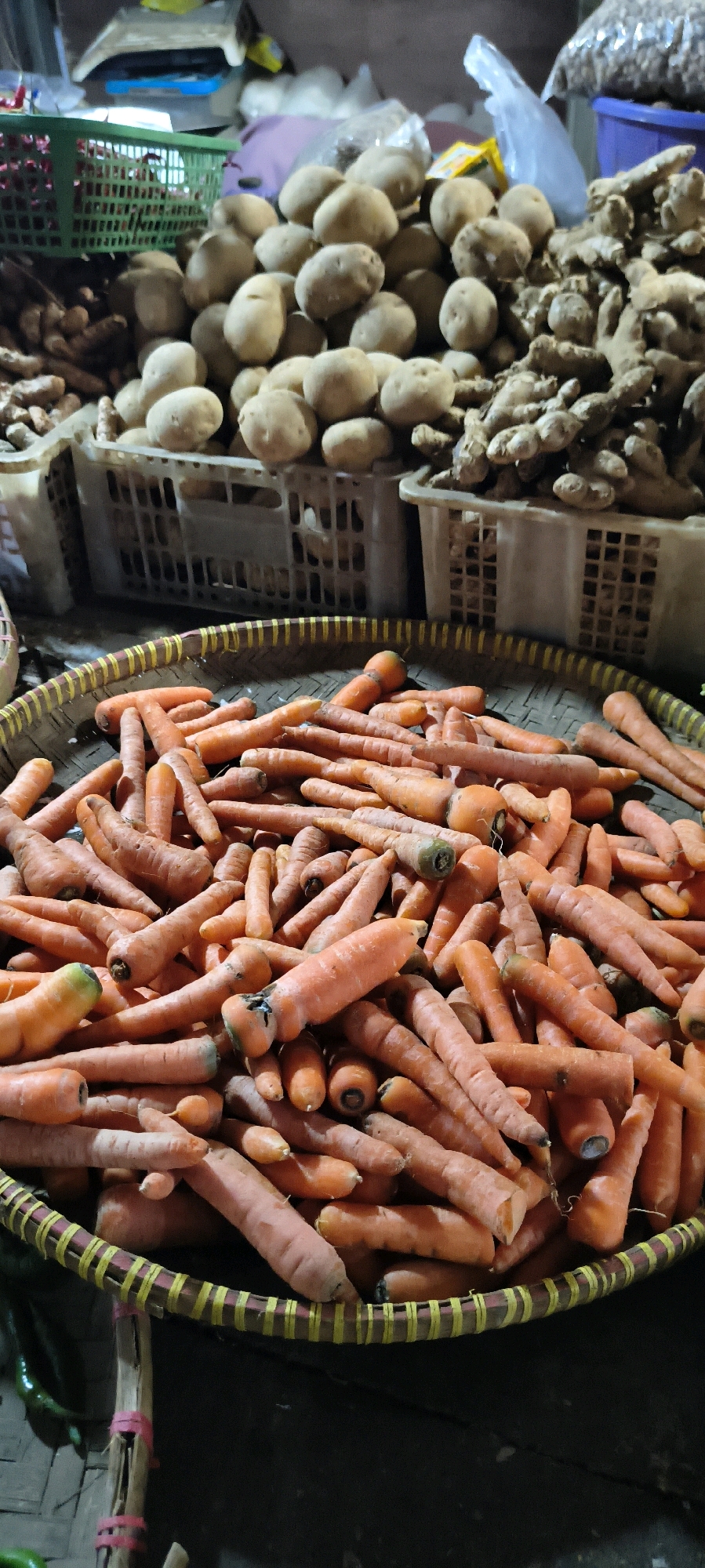 In Weru Lor, Indonesia, a woven basket filled with numerous fresh carrots is displayed at a market stall. The carrots are predominantly orange, with some showing hints of green near their tops. They are arranged in a dense pile, with their conical shapes and varying lengths clearly visible. The basket itself has a woven structure with yellow and red accents along its rim. In the background, other produce items, including potatoes and ginger, are visible in plastic crates, suggesting a vibrant market environment. The lighting in the scene is somewhat dim, indicating it could be early morning or late evening.