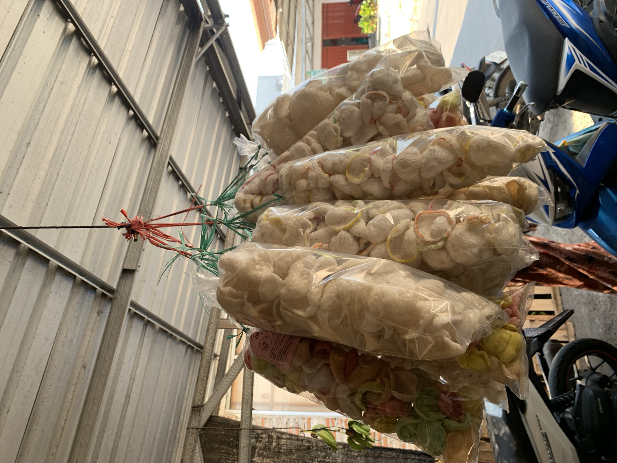 The image displays a vertical arrangement of numerous clear plastic bags, each filled with uncooked prawn crackers (kerupuk). These bags are bundled together with red and green strings and hung from a black rope or wire that stretches horizontally across the upper middle of the frame. The crackers are predominantly off-white or light beige, though some bags contain a mix of colorful crackers in pink, yellow, and green.
In the background on the left, a corrugated metal wall or fence is visible, showing a slightly weathered, industrial appearance. On the right side, parts of a blue and white motorcycle are partially visible in the upper background, with hints of another dark-colored motorcycle behind it. The scene is brightly lit by natural sunlight, indicating it is daytime with clear weather. No people are visible. A pink label on one of the lower bags has partially visible text "KERUPUK...", confirming the product. The overall setting suggests an outdoor environment, possibly a street vendor's setup, a market area, or a side alley.