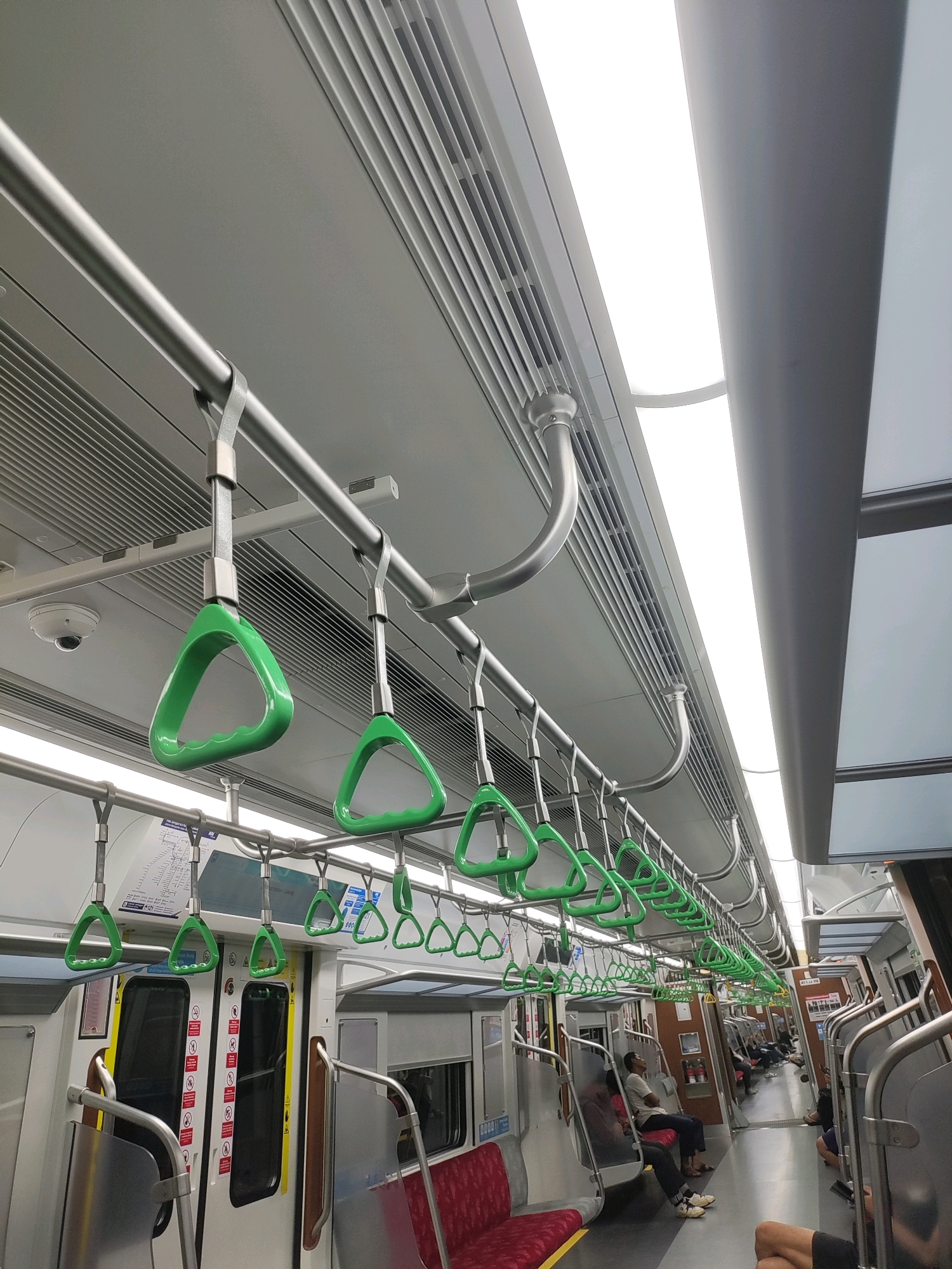 An interior view of a modern train carriage on the Jabodebek Light Rail Transit (LRT) in Bekasi, Indonesia. The carriage features a clean, metallic interior with bright fluorescent lighting panels overhead. Numerous green triangular grab handles are suspended from silver horizontal rails running along the ceiling. Metal poles provide additional support. The floor is a light gray. On the left side, there are white doors with various warning symbols displayed. Red seats are visible in the foreground, with some passengers seated in the middle and background of the carriage. The overall atmosphere is one of orderly transit.