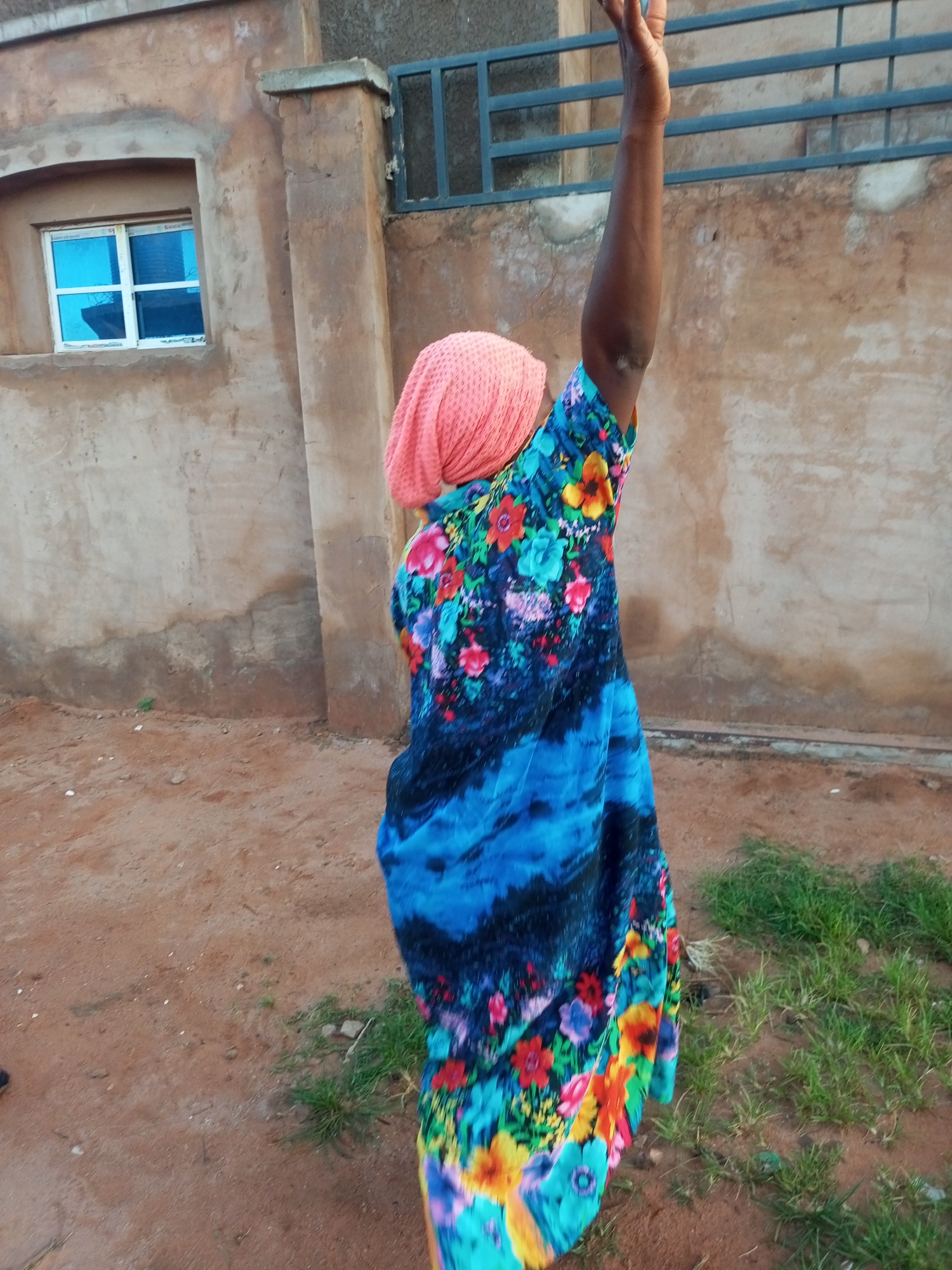 A person wearing a pink headscarf and a colorful floral dress is raising their right arm in the air. The person is standing on reddish-brown dirt ground with patches of green grass. Behind the person is a tan, textured wall with a small window that has blue glass. Further in the background, there is a dark metal railing. The scene appears to be outdoors, possibly in a residential area of Tempure, Nigeria.