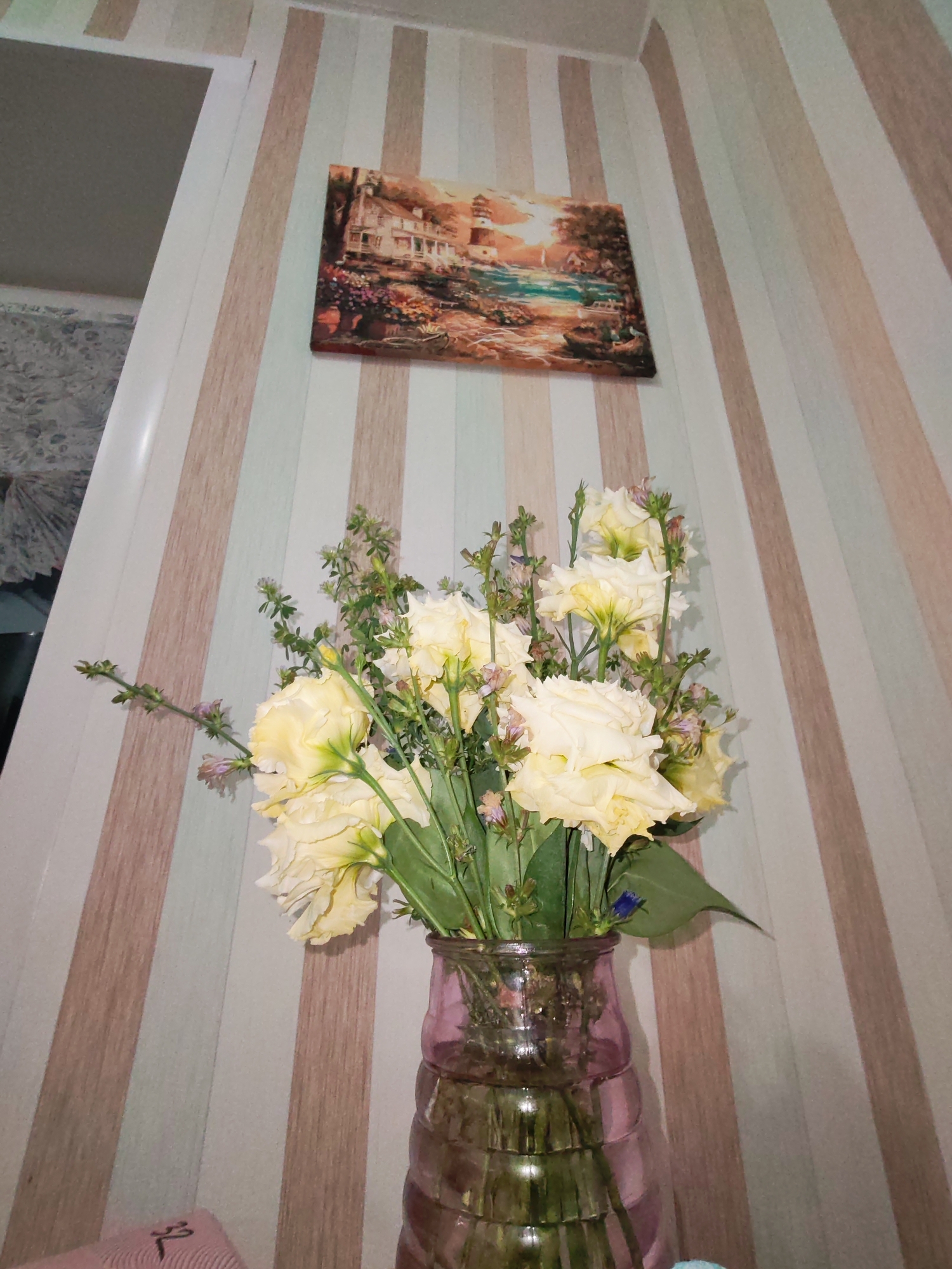 This image depicts a still life arrangement of yellow roses in a pink glass vase. The vase is positioned on a surface, likely a table, in front of a wall adorned with vertical striped wallpaper in alternating shades of beige and white. Above the floral arrangement, a framed painting hangs on the wall, featuring a landscape with a house and a lighthouse. To the left of the arrangement, a white vertical molding suggests a corner or doorway. The lighting appears to be ambient and likely artificial, casting soft shadows. The overall scene suggests an indoor setting, possibly a home interior. The provided image does not contain any information to determine its location within Izhevsk, Russia.