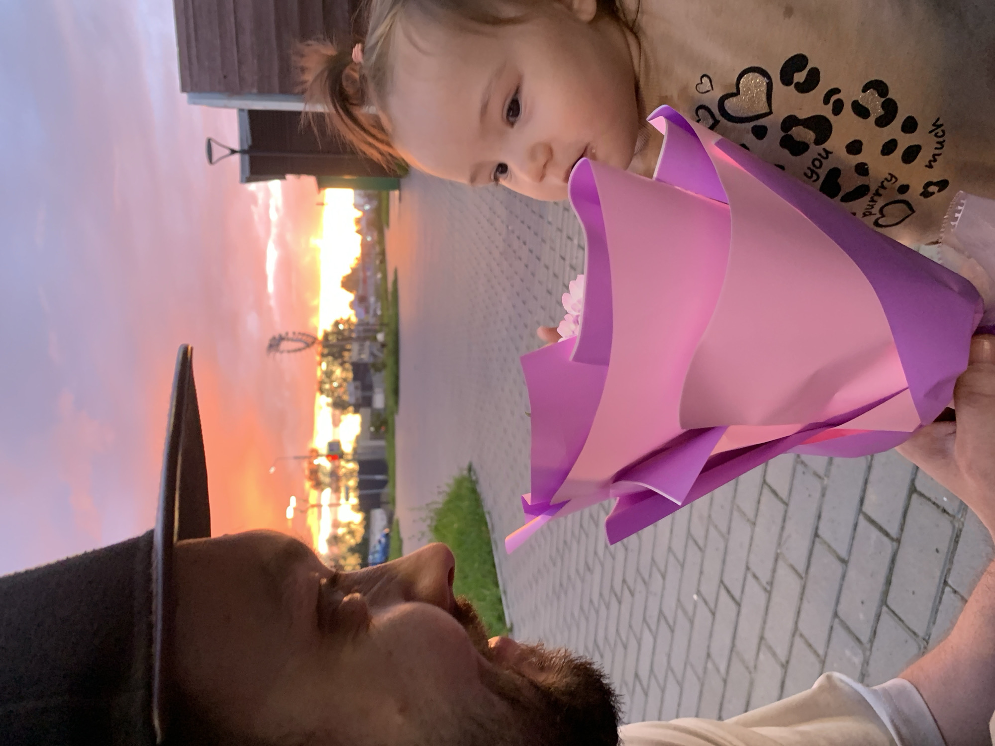In Chaikovsky, Russia, during sunset, a man wearing a hat is presenting a bouquet of pink and purple paper flowers to a young child. The man's face is partially visible, turned towards the child, with a smiling expression. The child, with hair tied in a ponytail, is looking at the bouquet and appears to be about to touch it. The child is wearing a light brown shirt with a leopard print and heart design, and text that reads "you purrry much". They are positioned outdoors on a paved walkway, with a grassy area to the left. In the background, a street with lampposts and buildings is visible under a colorful sky filled with clouds illuminated by the setting sun. A Ferris wheel-like structure is also visible in the distance.