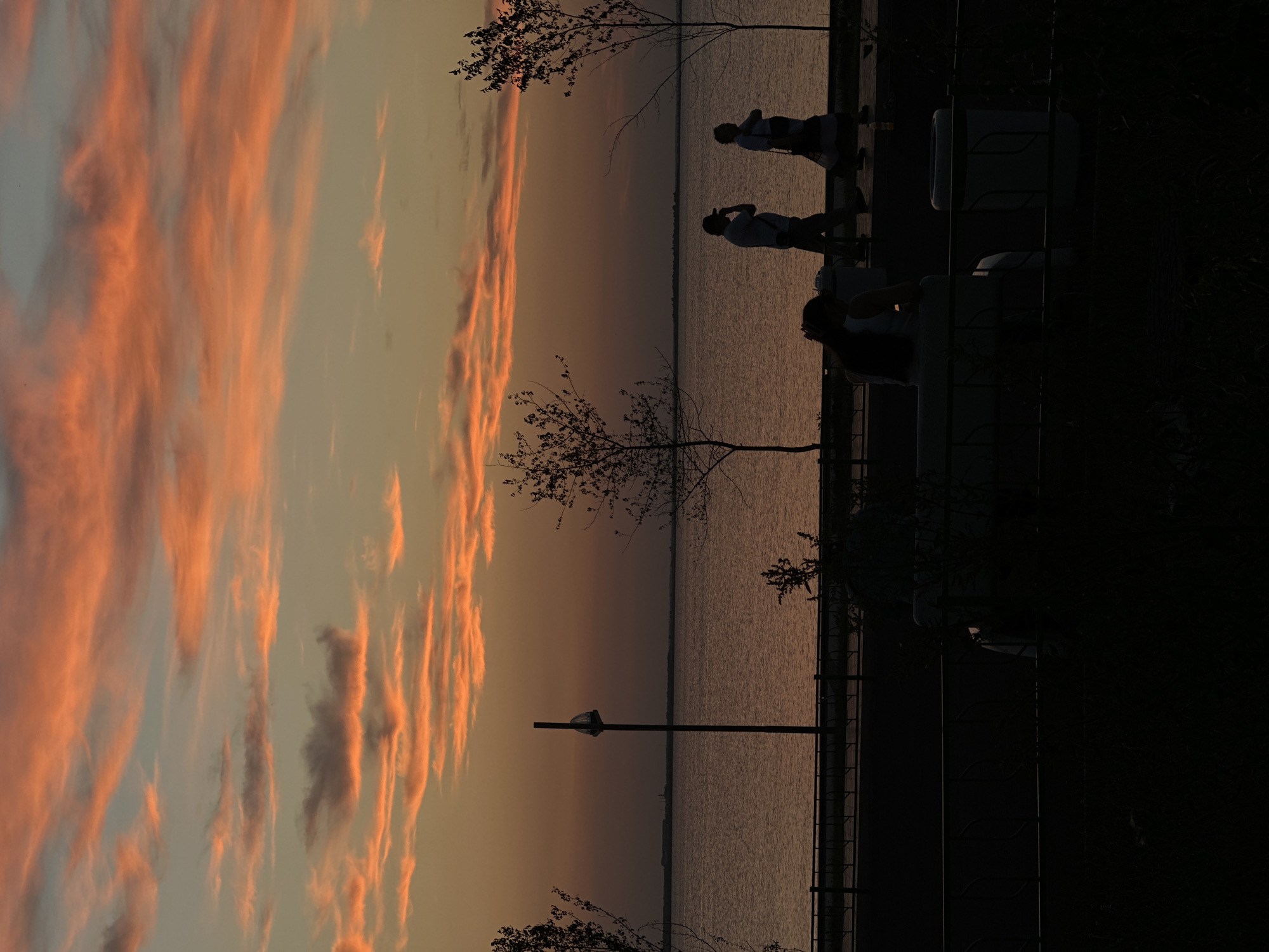 The image depicts a sunset over a body of water, likely a lake, on the outskirts of Kokshetau, Kazakhstan. The sky is filled with wispy clouds tinged with vibrant orange and pink hues, characteristic of a colorful sunset. In the foreground, silhouetted figures are visible. Two individuals are walking along a pathway next to the water, and another person is seated on a bench, facing away from the camera. Trees with sparse foliage frame the scene. A lamppost stands near the water's edge. The overall mood is serene, capturing the tranquil beauty of dusk in a natural setting.
