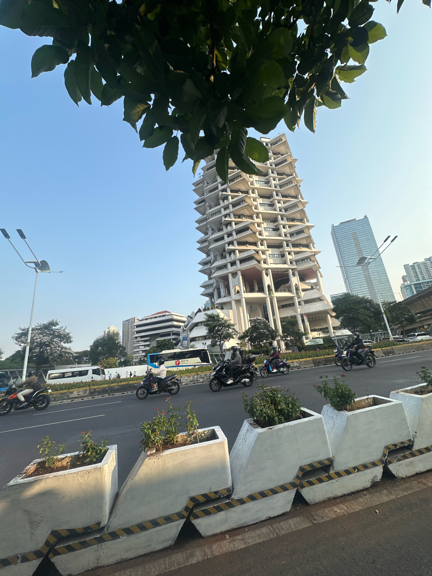 This is an eye-level, medium shot of a street scene in South Jakarta, Indonesia, captured under a clear blue sky. The prominent building in the background is the Wisma Nusantara, a distinctive modernist skyscraper with a unique stepped design and protruding balconies, likely from the 1970s. To its right, a more modern, glass-covered skyscraper stands tall.
In the foreground, a busy road is visible, with several motorcycles and a bus in motion. The motorcycles, some carrying riders and passengers, are traveling in the same direction. One bus, with "BRIN" written on its side, is also seen on the road.
The street is lined with concrete planters containing small green plants and red flowers. These planters are decorated with black and yellow hazard stripes along their bases. A tall street light with two lamps is positioned on the left side of the road. The upper portion of the image is dominated by the dark green leaves of a tree, providing a natural frame to the urban landscape. The overall impression is that of a bustling city environment with a mix of architectural styles and urban infrastructure.