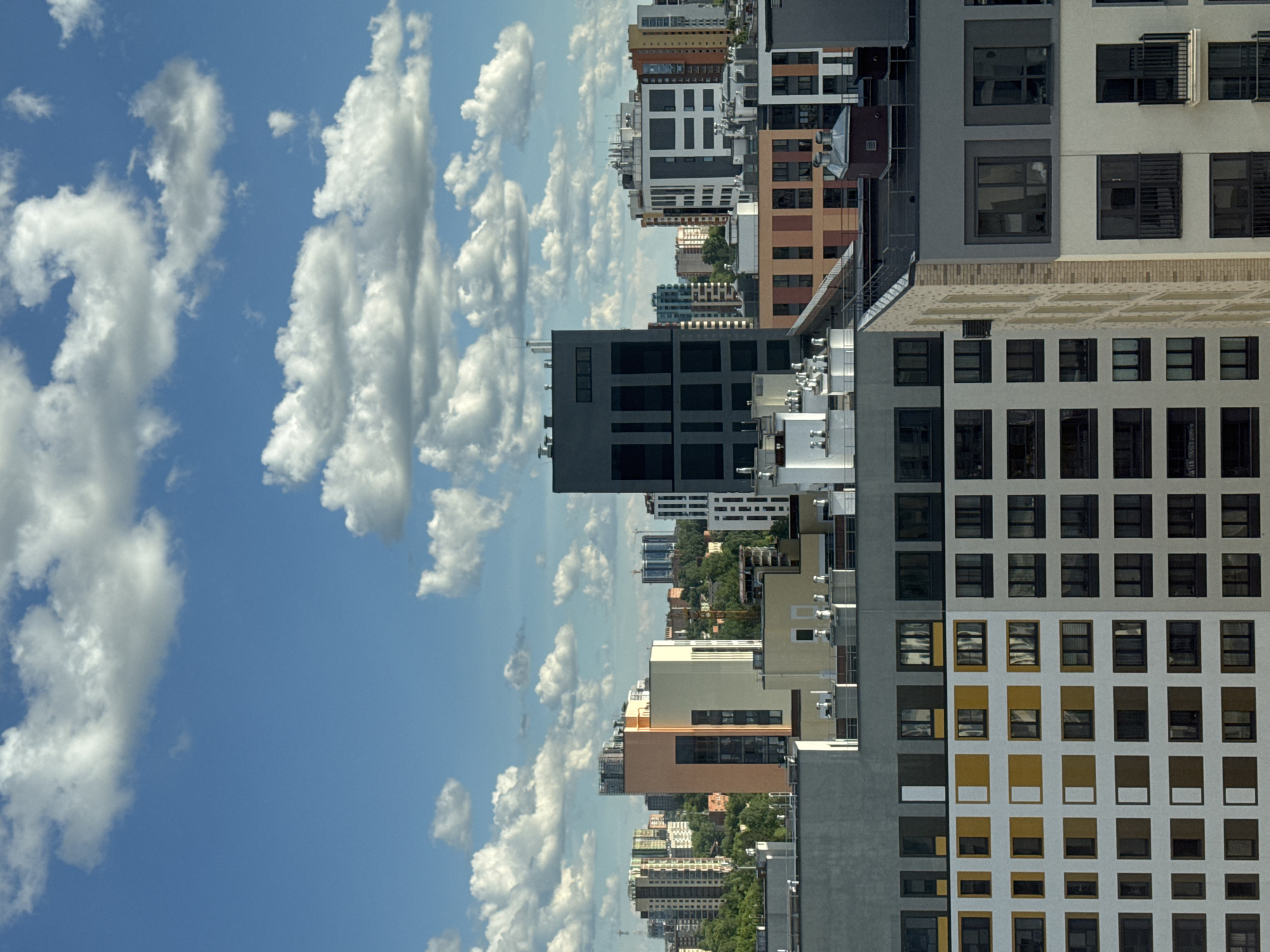 The image shows a cityscape of Izhevsk, Russia, viewed from an elevated position. The buildings are a mix of modern architectural styles, with varying heights, colors, and window designs. The sky is blue with scattered cumulus clouds. The perspective is angled, making the city appear vertical.