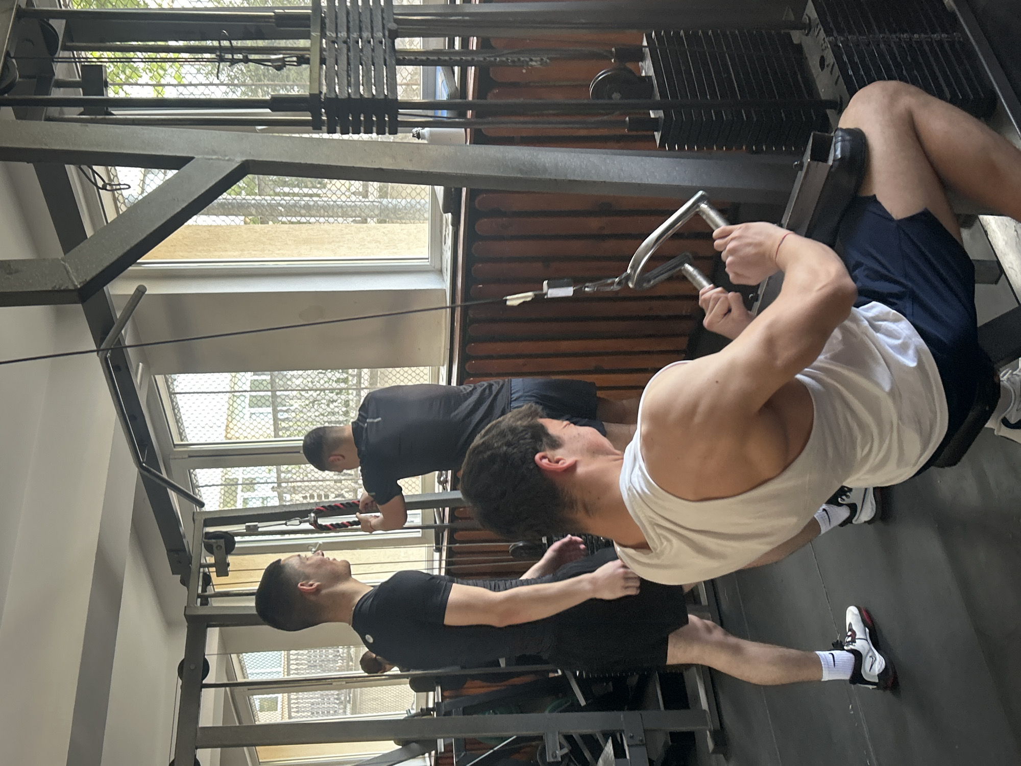 **Location:** Indoor gym, likely in Zlatograd, Bulgaria.
**People:** Three men are present, engaged in exercise.
**Subjects:** The men are the primary subjects.
**Objects:** Weight machine, weight stack, handles, gym equipment, clothing (white tank top, dark shorts, black shirt), footwear (sneakers), windows, metal framing, interior walls.
**Actions:** Men are using gym equipment, presumably for weight training or strength exercises. One man is gripping handles. Another man is using a machine that requires one hand to push.
**Interactions:** Men are actively engaged in exercises on the weight machine and other gym equipment.
**Environment:** The indoor environment is a gym setting with metal framing and equipment. Windows provide external light.