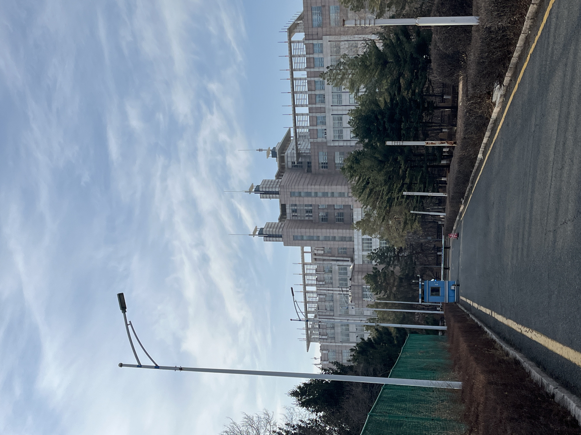 The image shows a street scene in Changchun City, China. A multi-story building with a distinctive stepped design is the main subject. It appears to be either a residential or office building. The sky is visible with wispy clouds. A street lamp stands on the left side of the road, which is marked with a yellow line. A guard booth and some greenery are visible along the edge of the road.