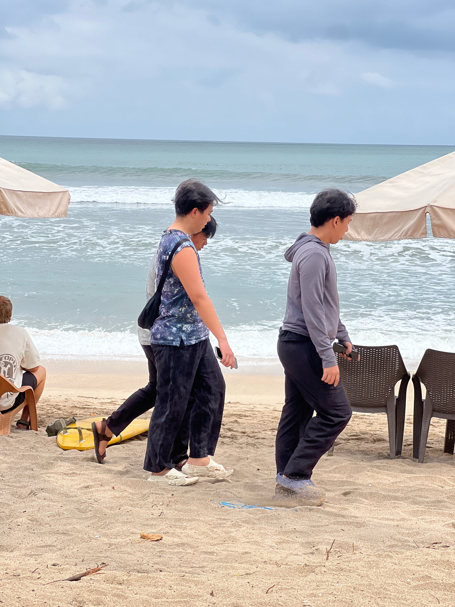 In Kuta, Indonesia, three individuals are walking on a sandy beach. The person in the foreground on the right wears a grey hooded long-sleeved shirt and black pants, holding a black mobile phone. To their left, another person in a blue and black patterned sleeveless shirt and black pants walks ahead. A third person is partially visible behind them, wearing a grey shirt. In the background, the ocean with moderate waves stretches to the horizon under a cloudy sky. Two large, light-colored umbrellas are set up on the beach, and some dark plastic chairs are visible. A yellow flotation device lies on the sand near the two walking figures. A person with light-colored hair sits on a chair to the far left of the image, facing away from the camera.