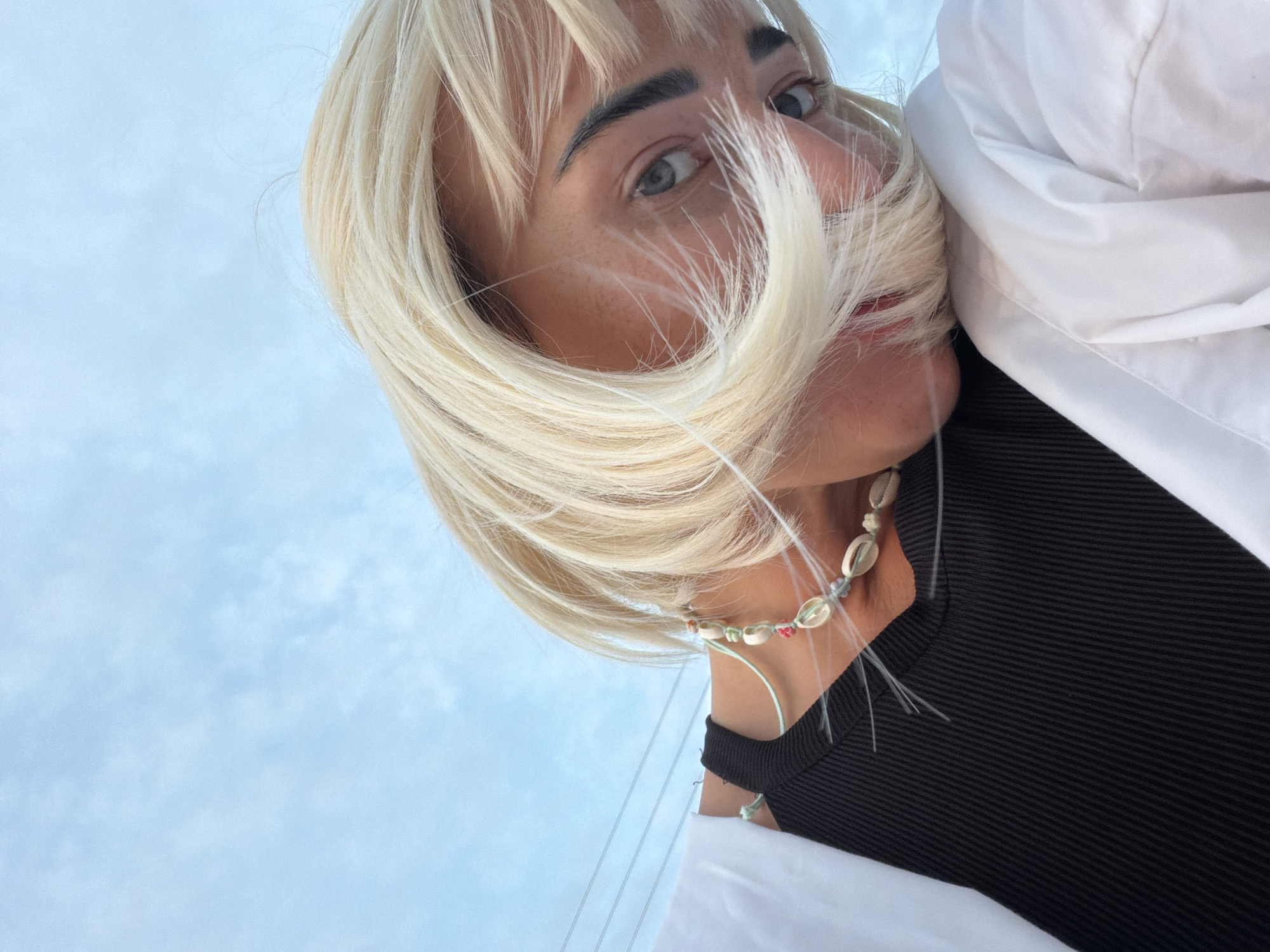 A young woman with blonde hair, wearing a black tank top and a white open shirt, is captured in a close-up, low-angle shot against a sky of pale blue with wispy clouds. Her face is tilted downwards, with her blonde hair obscuring part of her face and resting on her shoulder, which is covered by the white shirt. She is wearing a necklace made of small shells and beads. Her left eye is visible, looking towards the camera. The image appears to have been taken outdoors, possibly in Sochi, Russia, given the clear sky and the presence of what look like power lines in the background. The lighting suggests it is daytime.