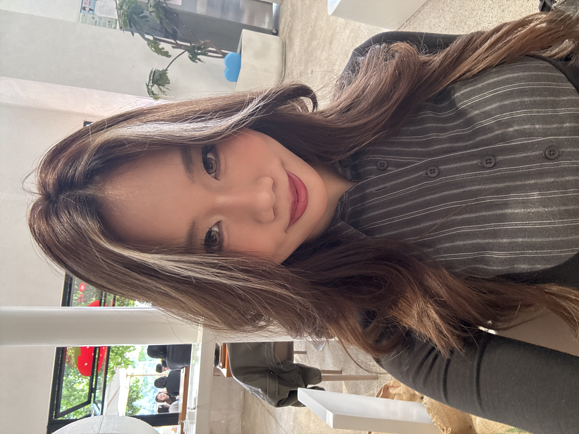 A woman with brown and blonde highlights poses indoors. She wears a gray striped button-up shirt. Outside the window, there are trees and what appears to be other people. Given the location information, it is likely this image was taken in 江北城街道, China.