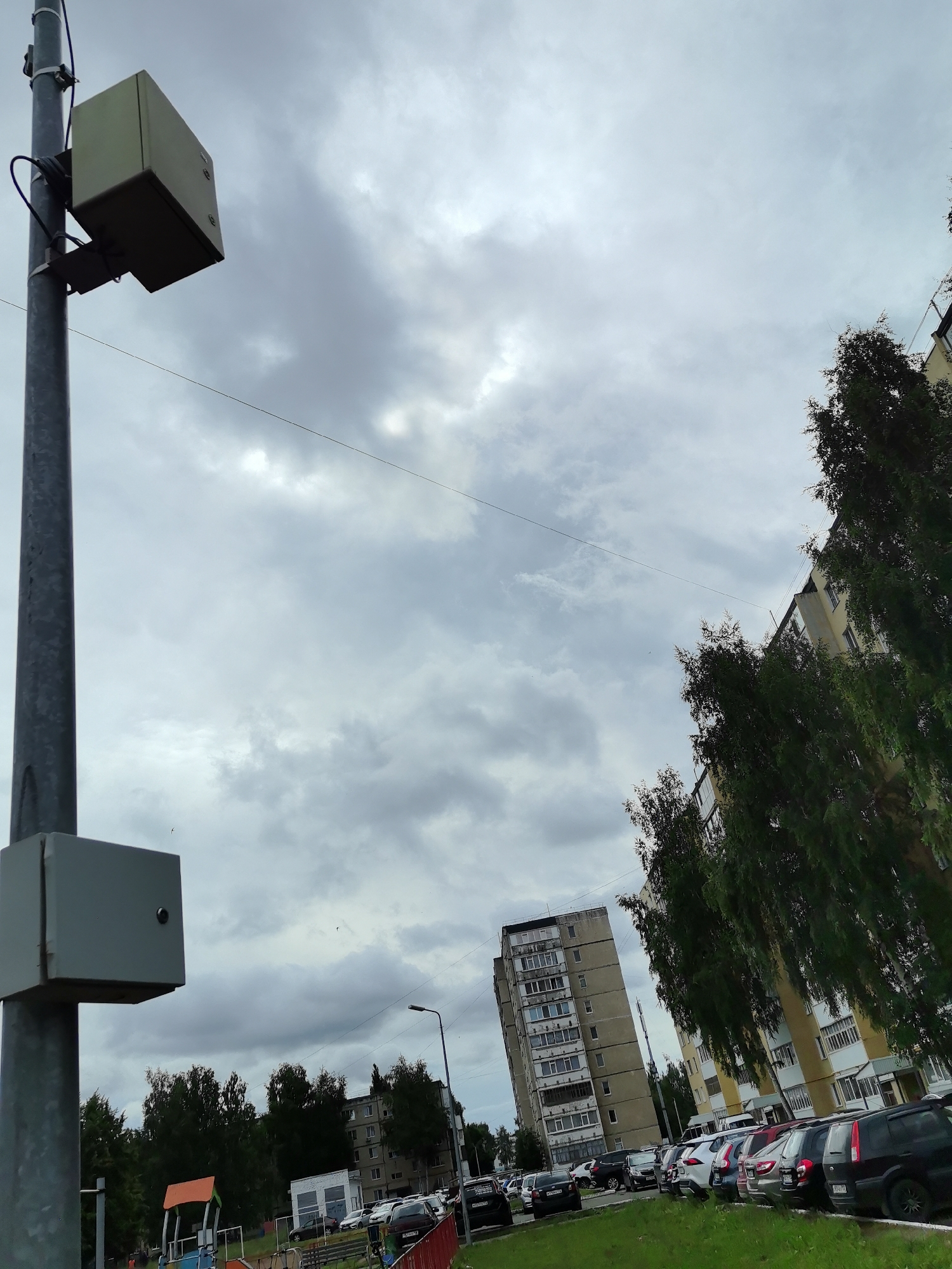The image shows a city street scene in Neftekamsk, Russia, on a cloudy day. In the foreground, a metal pole with two gray boxes mounted on it stands on the left side of the frame. The upper box appears to be a camera or surveillance device, while the lower box is a larger, rectangular enclosure. A children's playground with a red and orange structure is partially visible behind the pole. A row of trees lines the middle ground, separating the playground from a parking lot filled with various cars. Beyond the parking lot, two apartment buildings are visible. The building on the left is a tall, concrete structure, while the building on the right is a lighter-colored building with yellow accents. The sky is overcast with gray clouds. No people are visible in the frame.