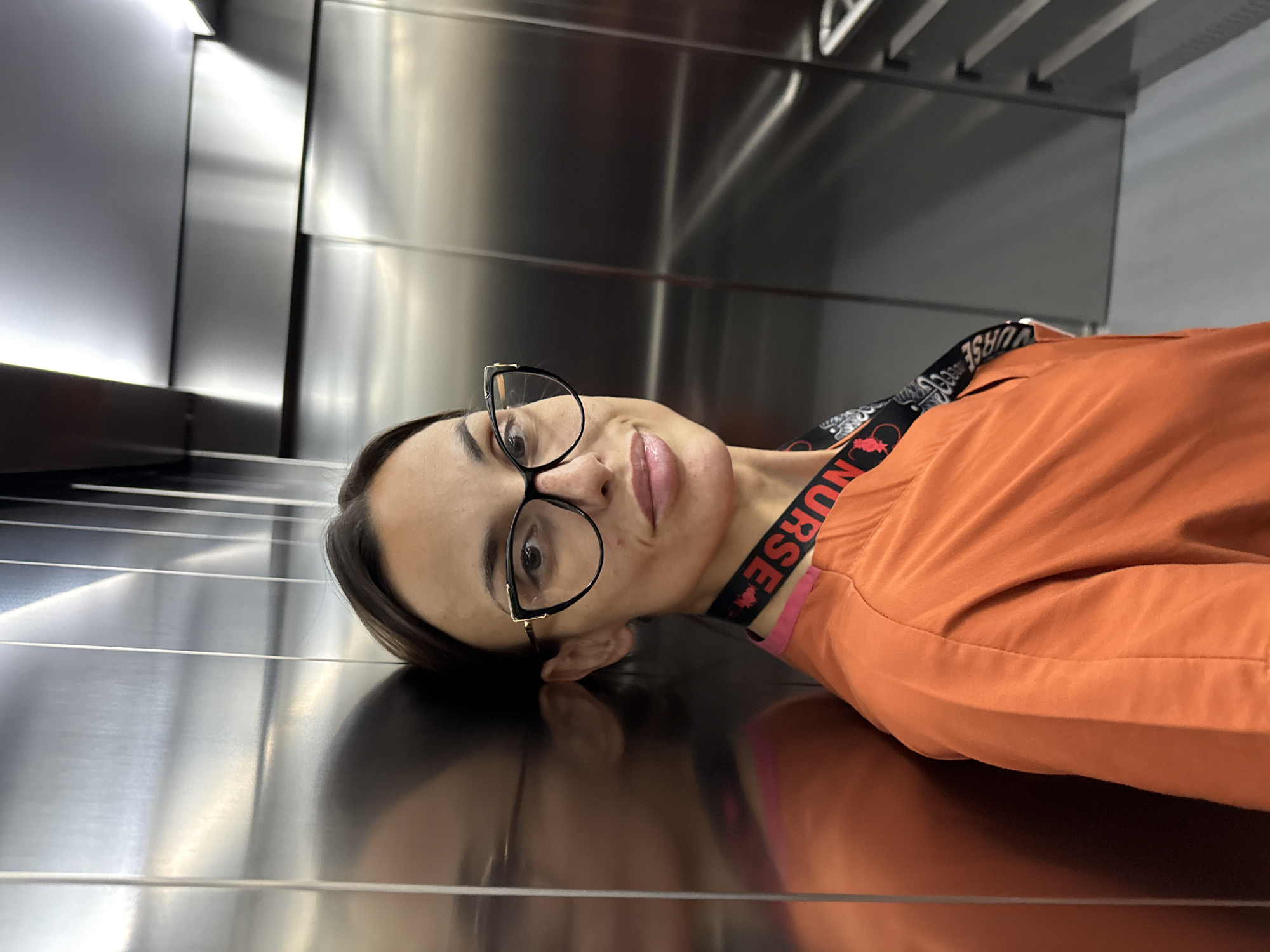 A woman with dark hair pulled back and wearing black-rimmed glasses, a bright orange scrub top, and a lanyard with the word "NURSE" printed on it, poses for a selfie inside an elevator with stainless steel walls. She is leaning against the left wall of the elevator, with her reflection visible. The elevator appears to be in a modern building. The specific location in Pleven, Bulgaria is not discernable from the image itself.