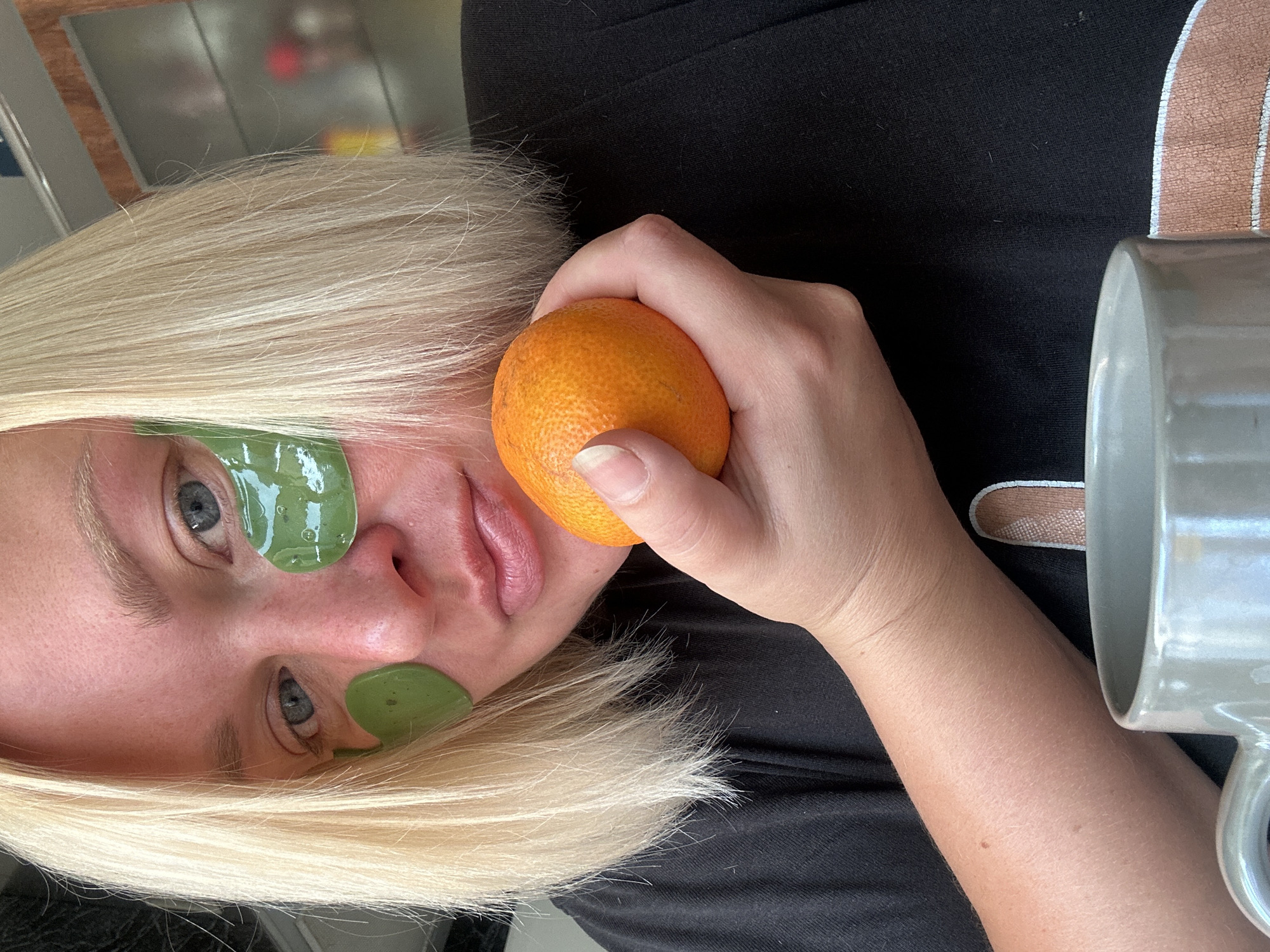 A close-up, waist-up shot shows a person with blonde hair and blue eyes, wearing under-eye masks and a black t-shirt. The person is holding a small orange fruit in their right hand, near their mouth. A grey mug is visible in the foreground. The background is blurred, showing some shelves with objects. There is no indication of the location being Chaikovsky, Russia.