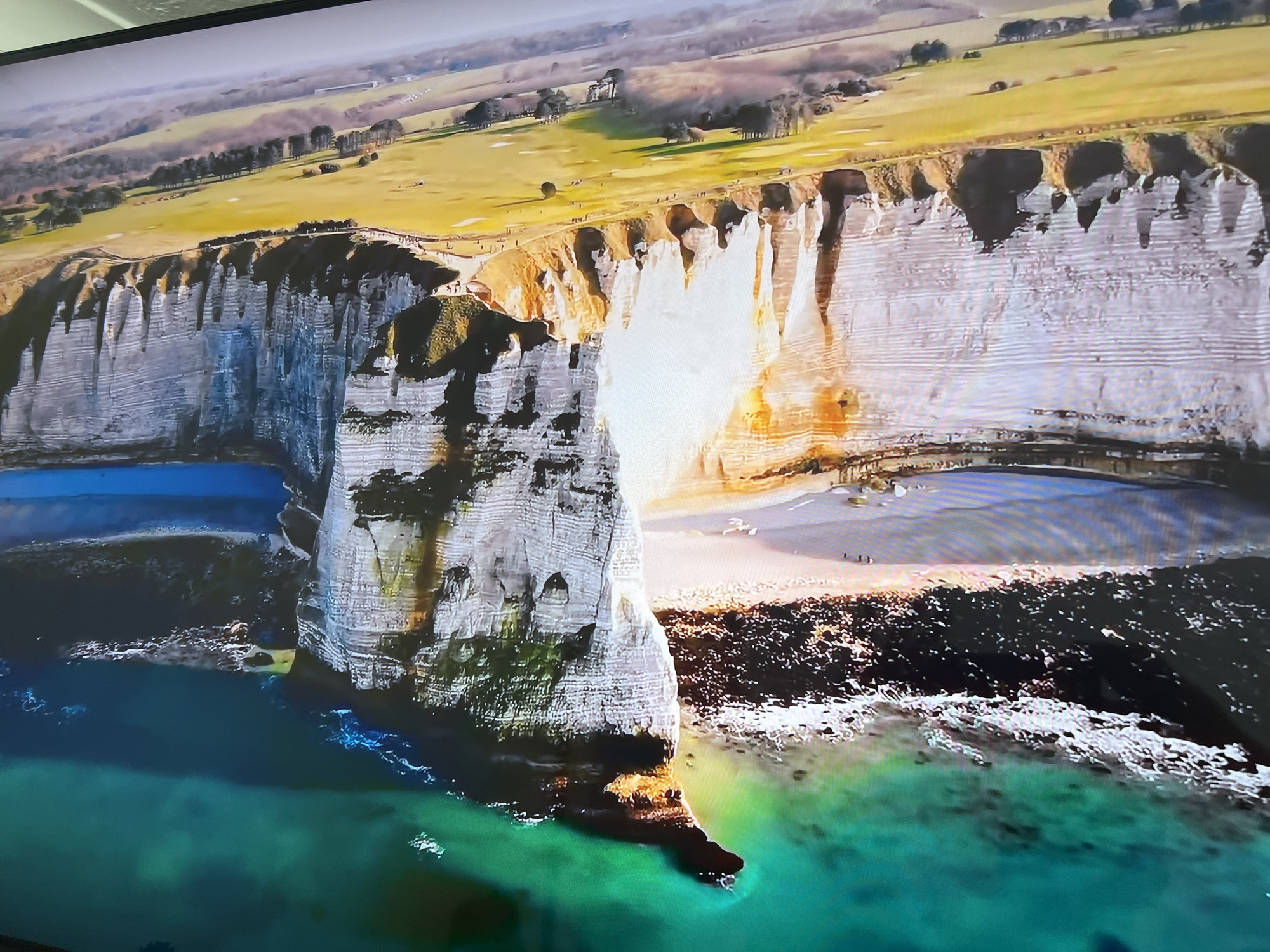The image shows a coastal landscape, specifically the cliffs of Étretat in Normandy, France, not Basingstoke and Deane, United Kingdom. The prominent feature is a large, natural chalk arch formation extending into the sea, partially covered in vegetation. The water near the cliffs is turquoise in color. Above the cliffs, there is a grassy plateau with a golf course. A small beach is visible at the base of the cliffs with a few people on it. The top of the cliffs appear to have people walking along them.