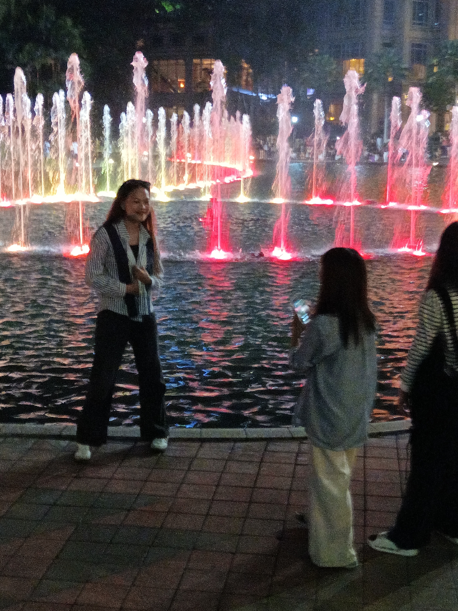 The image depicts a nighttime scene at a brightly lit fountain in Kuala Lumpur, Malaysia. In the foreground, three individuals are visible. A woman stands to the left, wearing a striped collared shirt over a dark garment and black pants. To her right, a person is holding a phone, facing away from the camera. Another individual, partially visible on the far right, wears a striped top and dark pants. The background features a large fountain with water jets illuminated by colorful lights, creating a vibrant display against the dark night sky and surrounding buildings. The ground is paved with interlocking tiles, and the water in the fountain reflects the colorful lights.