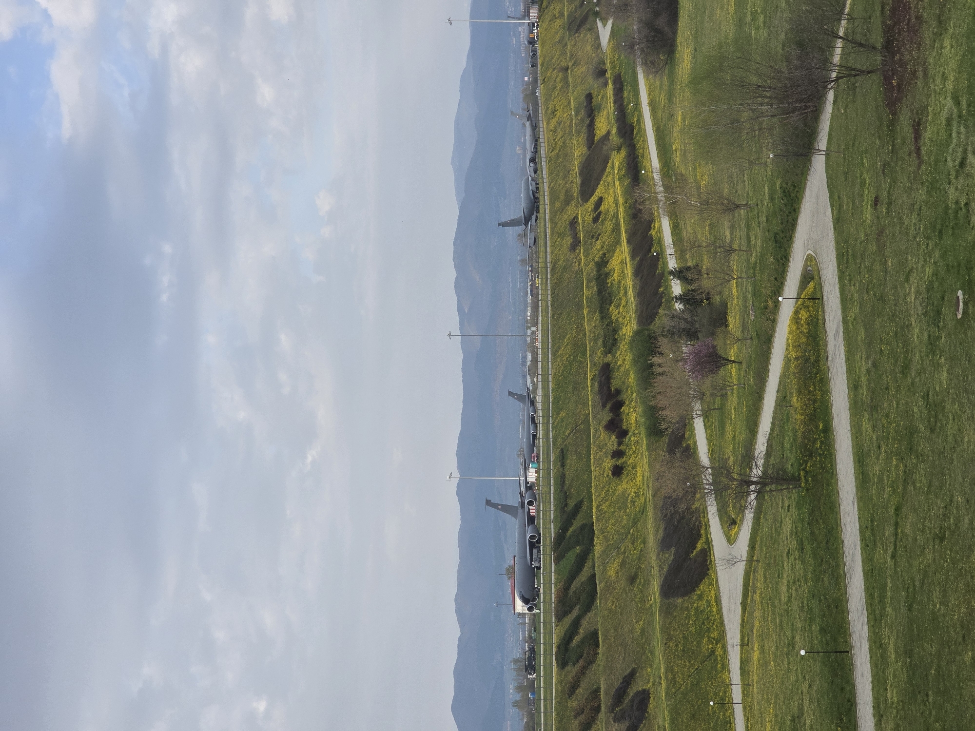 The image shows a park scene with airplanes in the background, located in Sofia, Bulgaria. The right side of the image is a grassy area with yellow flowers and walking paths. On the left, there is a view of mountains and a line of parked military aircraft along a runway. The sky is overcast.