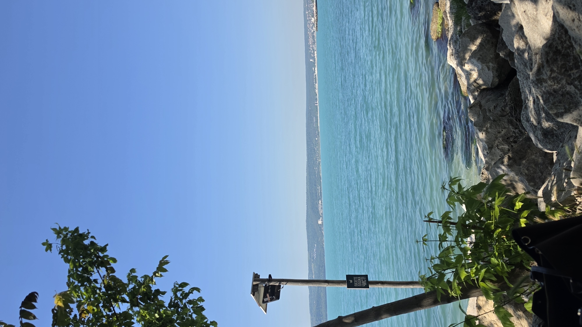 The image shows a view of the Black Sea in Varna, Bulgaria, from a vantage point close to the shore. The water is a light turquoise color, and the coastline is visible in the distance. On the right side of the image, there are large rocks and some greenery. A wooden pole with a sign on it is also visible, indicating a possible public area. The sky is a clear blue with a slight gradient towards the horizon. The angle suggests the photograph was taken with the camera oriented vertically.