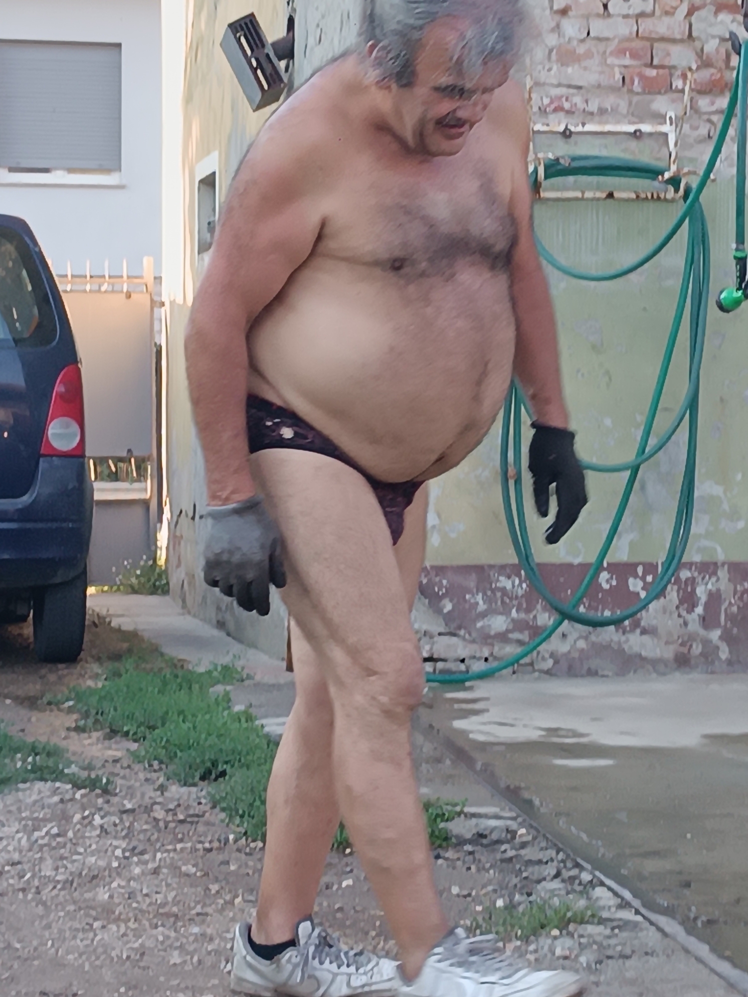 A shirtless, middle-aged man with graying hair and a mustache stands outside. He is wearing dark briefs, grey work gloves on his left hand, and black gloves on his right hand. He is also wearing white sneakers. A dark blue car is parked to his left, and a green garden hose hangs on the wall behind him. The man appears to be looking down and to his right. The scene takes place in what appears to be a residential area, possibly Viadana, Italy, with a building and a paved area visible.