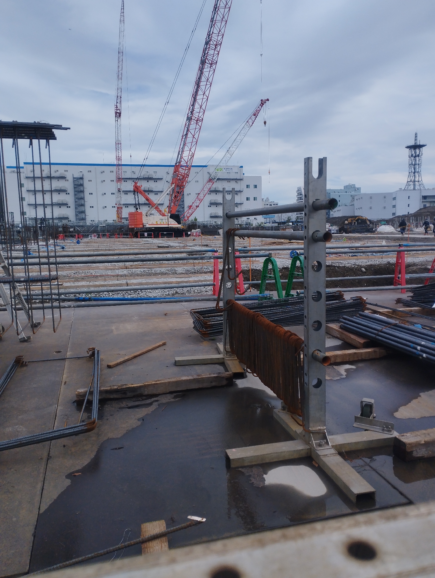 The image shows a construction site in Ichikawa, Japan, under an overcast sky. Dominating the scene are several large construction cranes, including a tall lattice boom crane and a larger, red and white mobile crane. In the background, a long, multi-story white building with numerous windows is visible, with a smaller, more industrial-looking structure behind it. The foreground is filled with construction materials and equipment, including rebar bundles, metal frames, wooden beams, and what appears to be pipes or conduits. Several red and green construction barriers are also present. The ground is a mix of concrete and dirt, with puddles of water suggesting recent rain. There are a few figures of construction workers visible in the midground, moving around the site, and a yellow excavator in the distance.