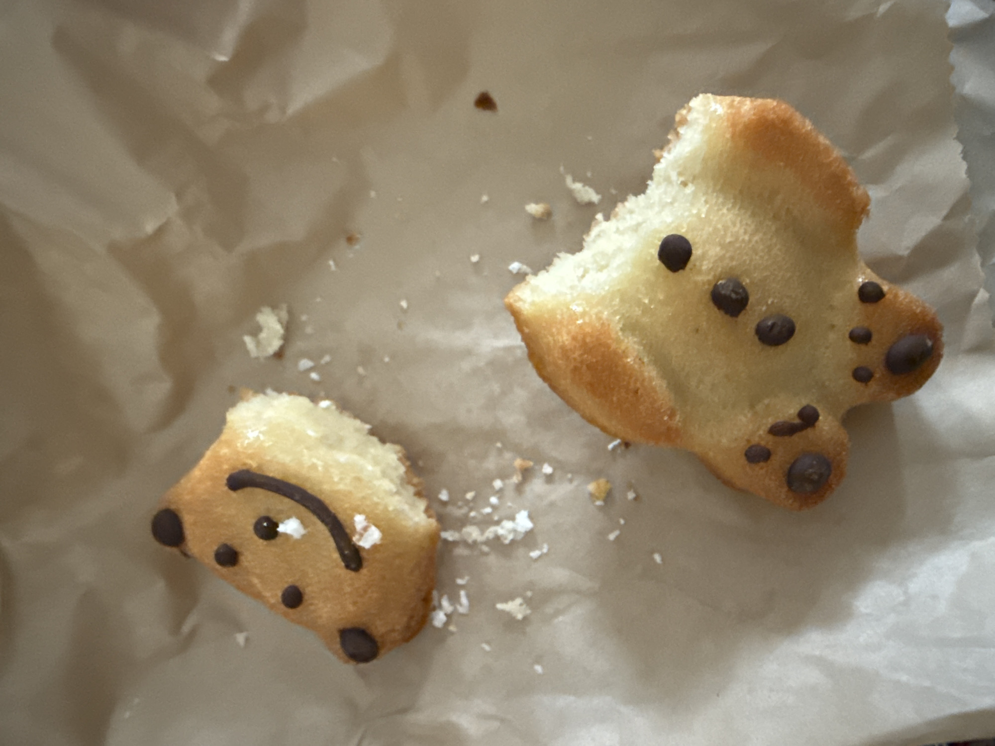 The image shows two broken animal-shaped pastries on a crumpled piece of parchment paper. The pastries are golden brown and decorated with dark chocolate chips, some of which are arranged to resemble faces. Crumbs are scattered around the pastries on the paper. The location is indeterminable.